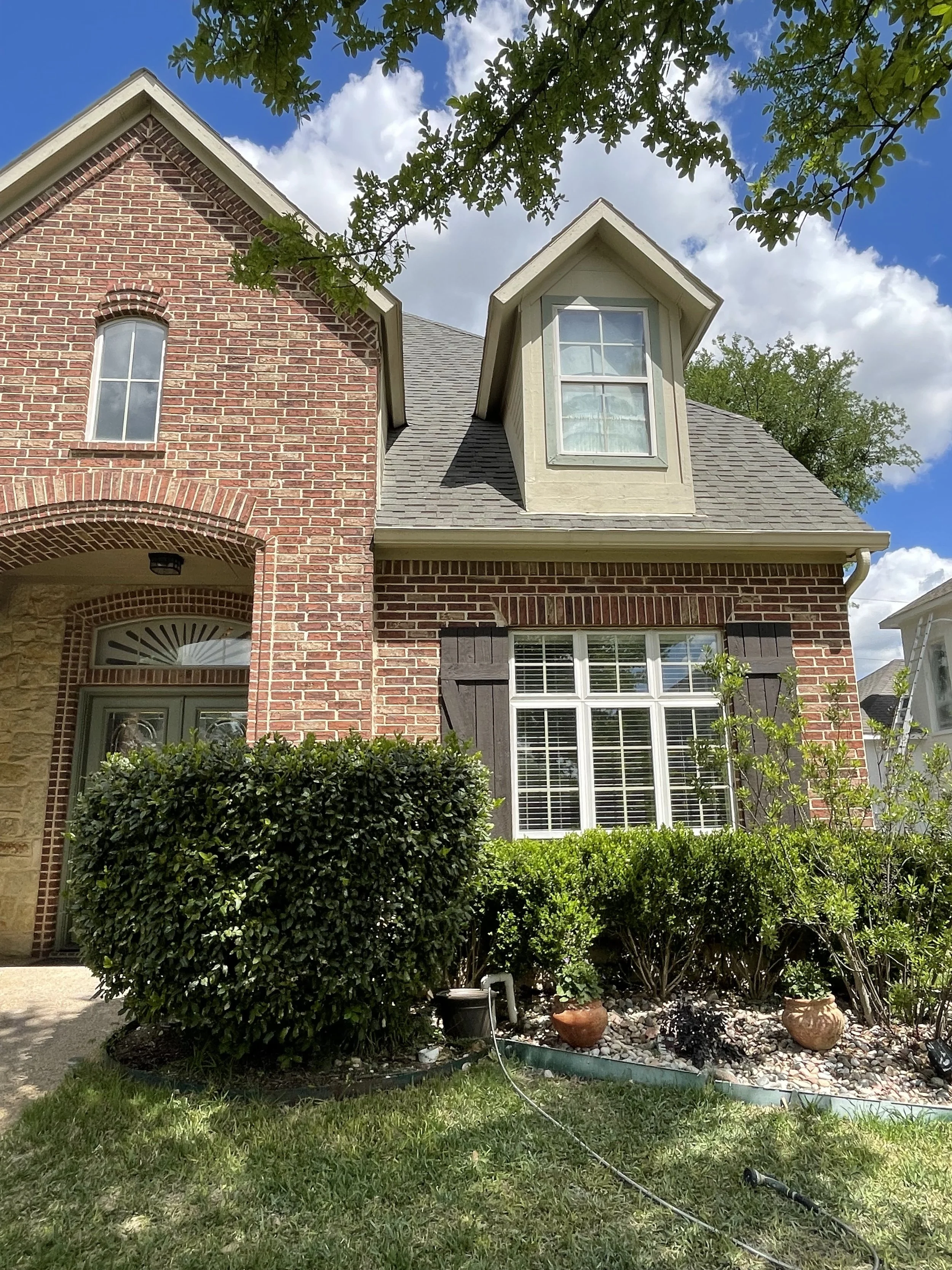 Front view of a brick house with a gabled roof, a window in the dormer, and a well-maintained garden in the foreground with shrubs, houseplants, and a lawn sprinkler on a sunny day with partly cloudy sky.