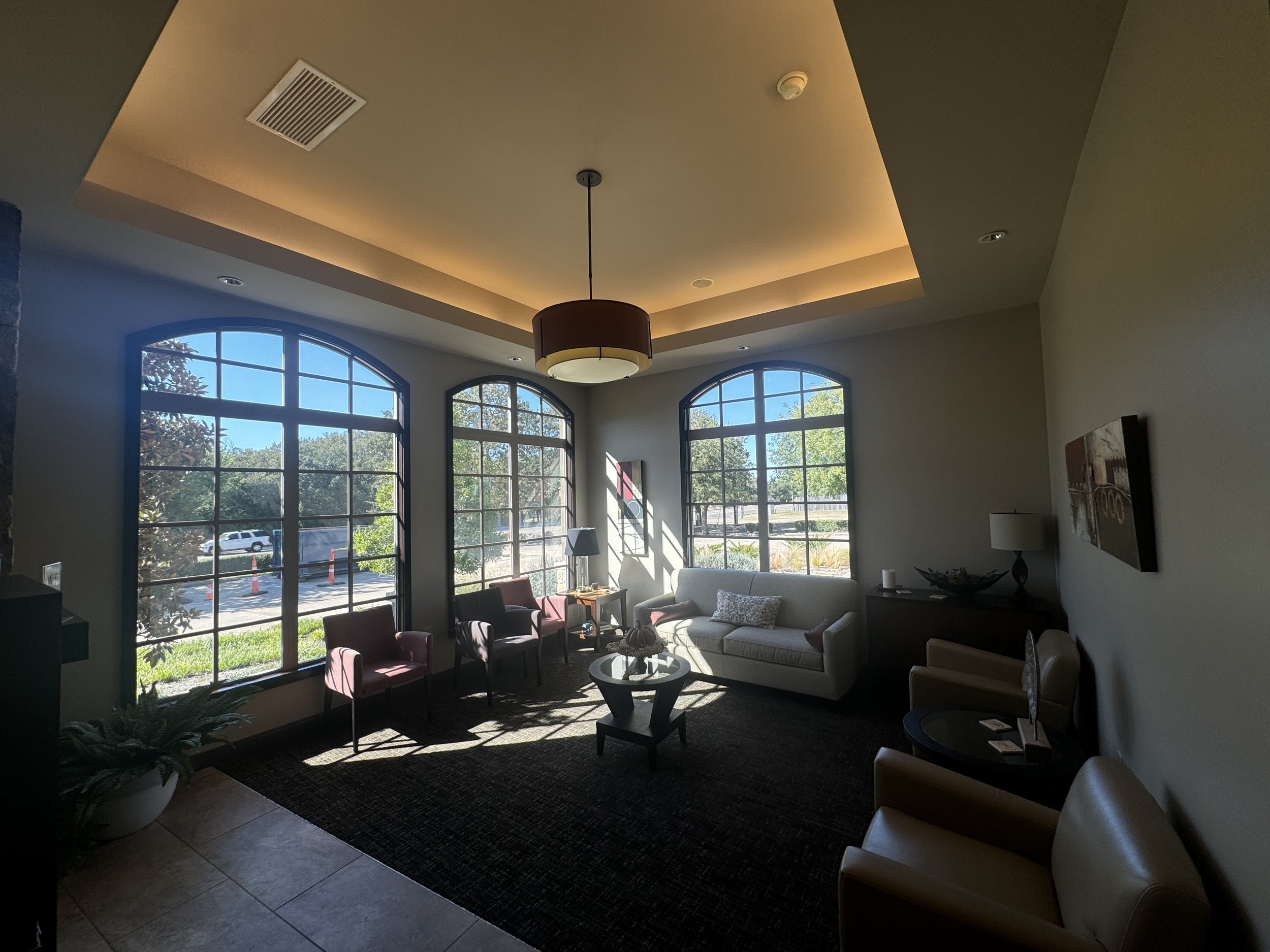 Indoor lounge area with large arched windows, cream-colored sofas, pink armchairs, a wooden coffee table, and a decorative bowl filled with plants.