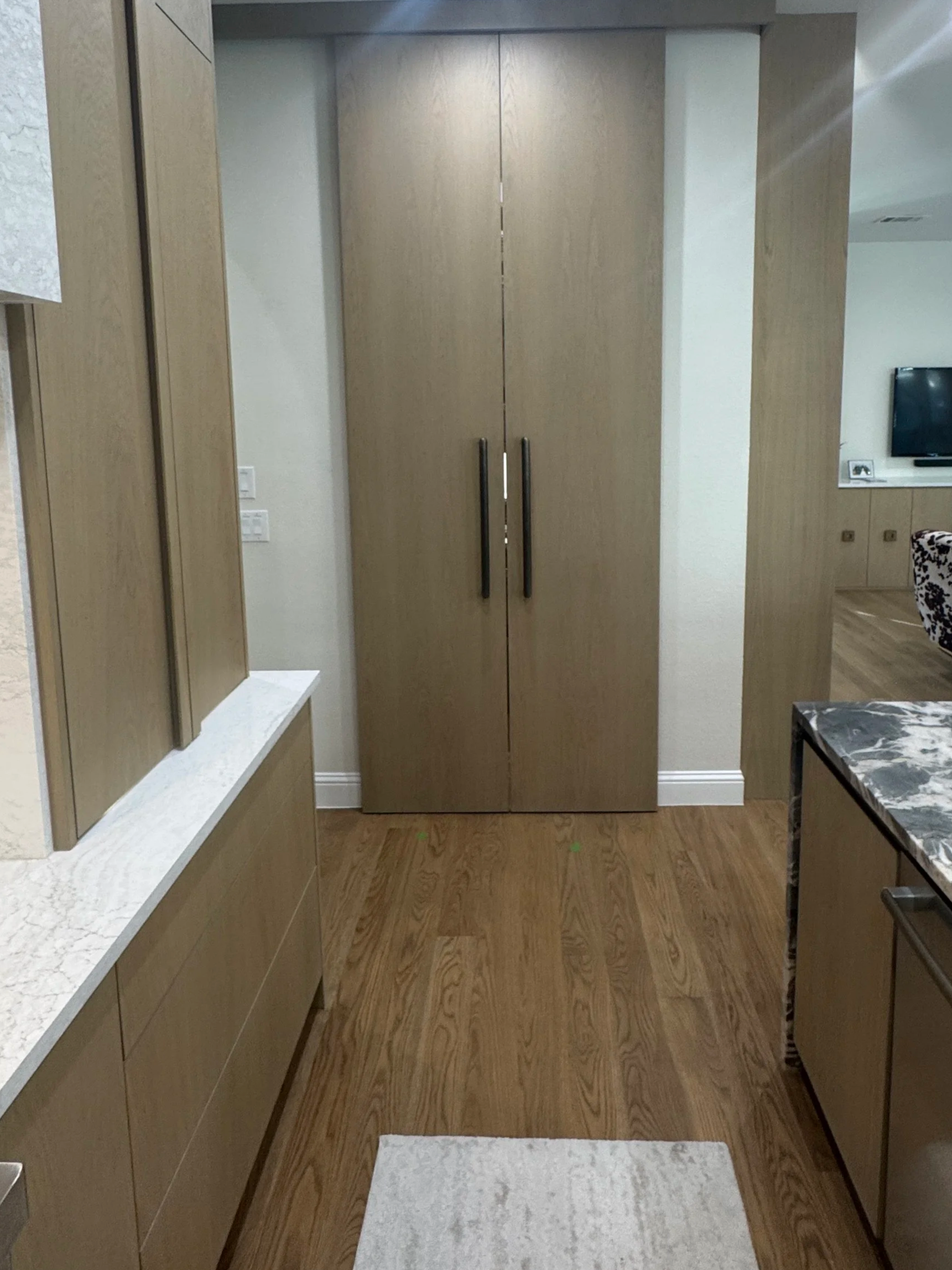 A hallway view with wooden floor and a large wooden cabinet with two handles in the center of the image. To the right, there is a partial view of a marble-topped table and a television on the wall in the background.