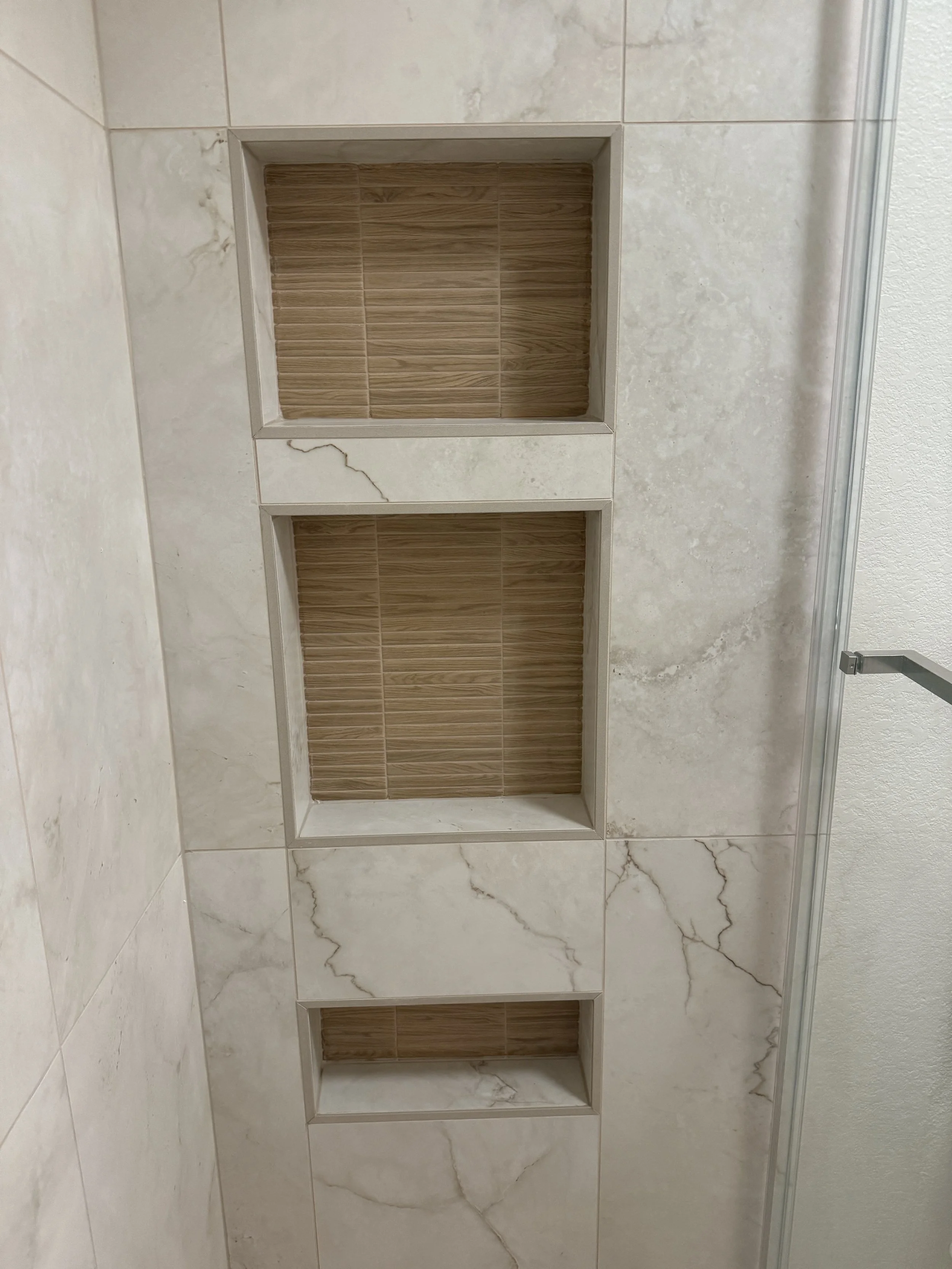 Three open wall tiles with a beige marble pattern, revealing recessed shelves with brown wooden back panels, in a bathroom wall.
