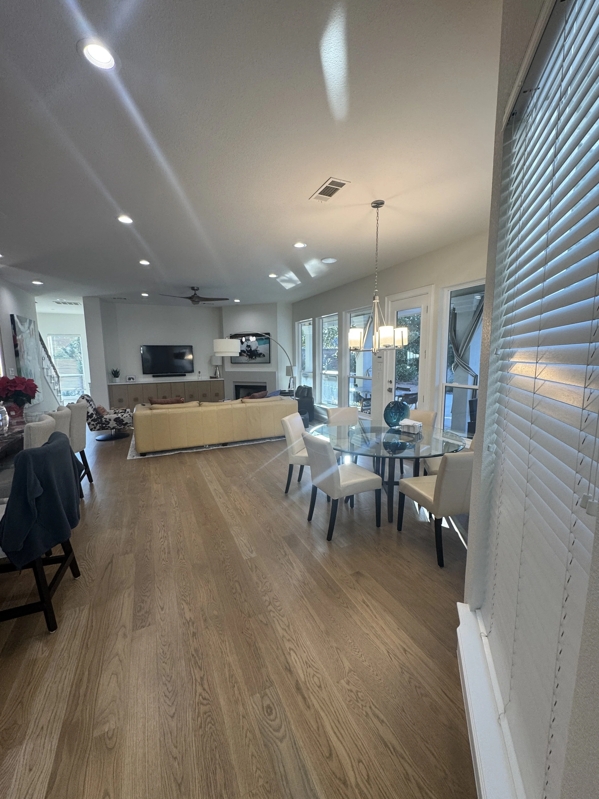 A spacious living room with wooden flooring, multiple white chairs, a beige couch, and sliding glass doors leading to an outdoor area. The ceiling has recessed lighting and a hanging chandelier. There's a TV mounted on the wall, and large windows with blinds letting in natural light.