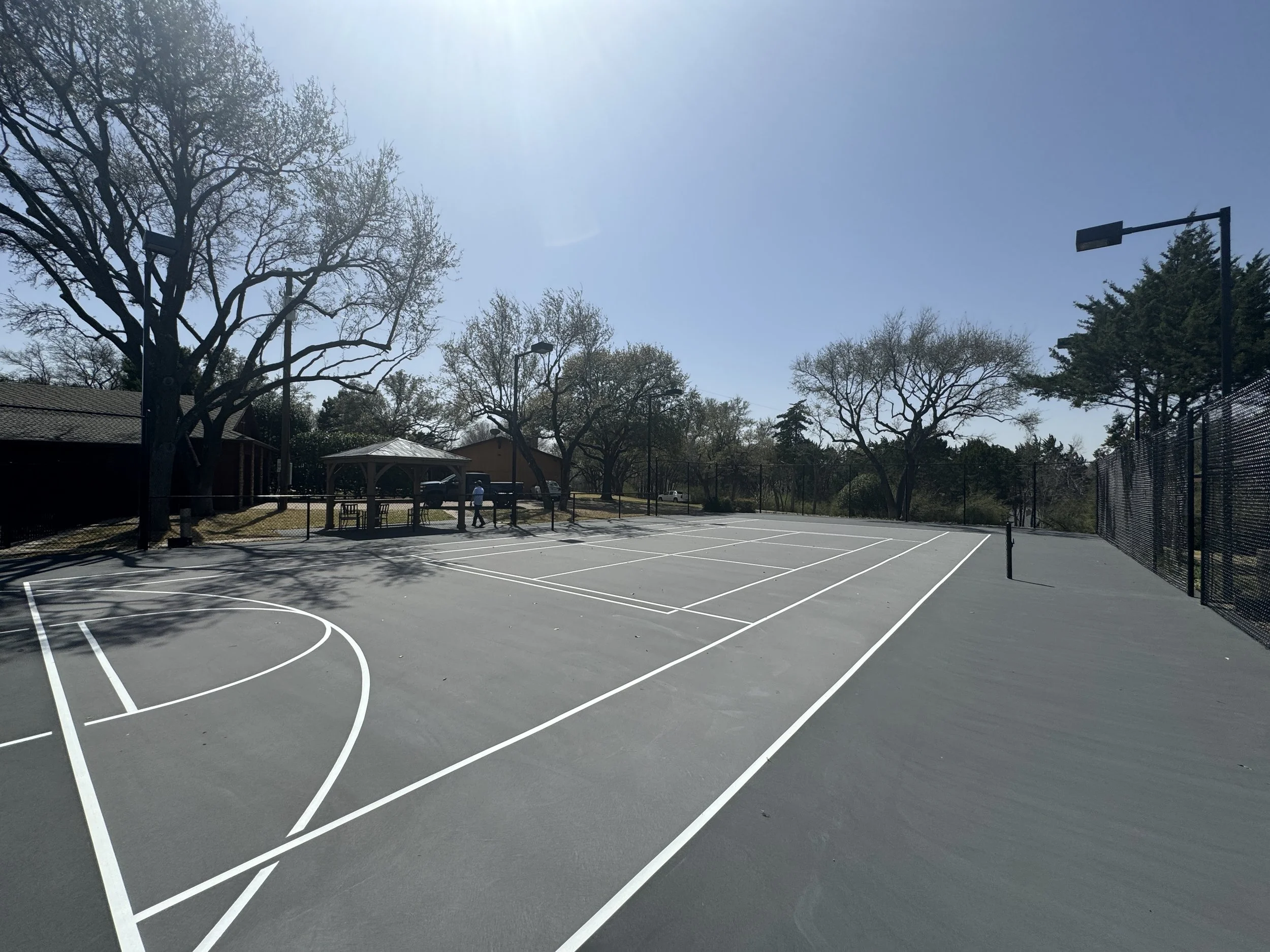 An outdoor basketball court with white painted lines and a chain-link fence, surrounded by tall trees with some leaves, a small building with a gabled roof, and metal lighting poles under a clear blue sky.