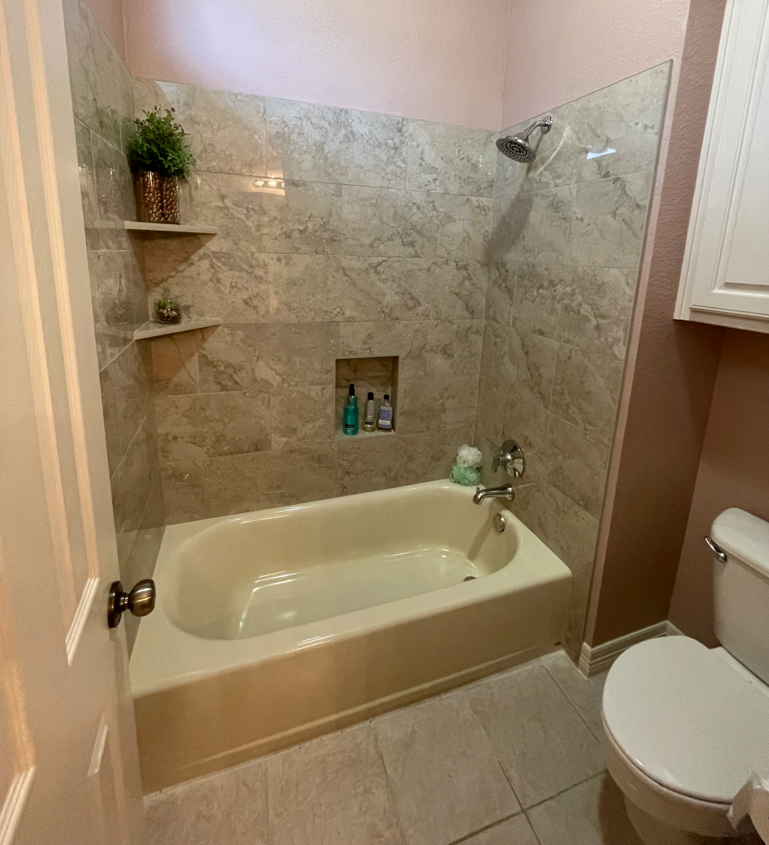 A bathroom shower area with beige marble tiles, a showerhead, and built-in shelving holding various bottles and a potted plant. A bathtub is visible in the foreground, and part of a toilet is seen to the right.