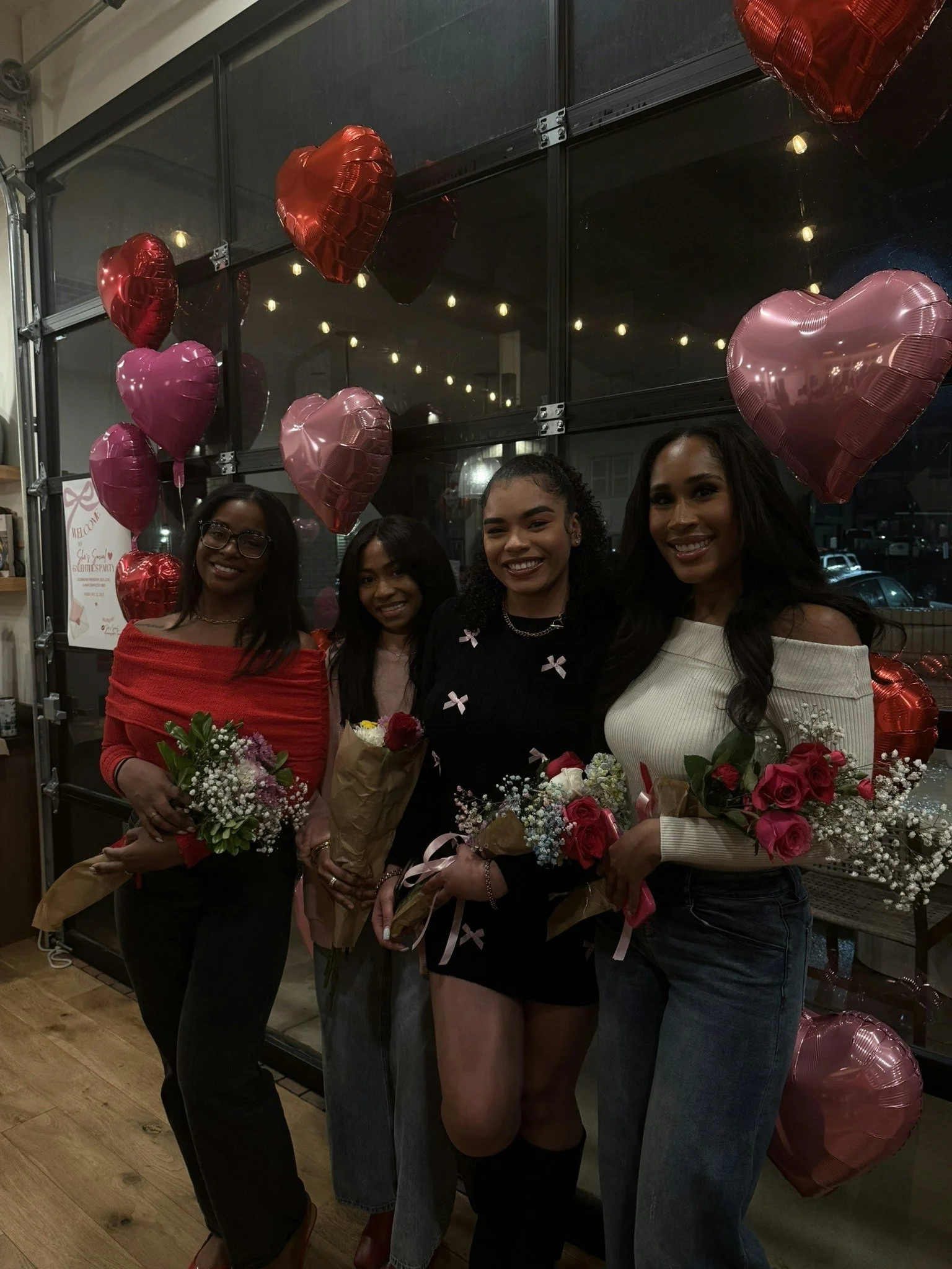 Four women holding bouquets of flowers, standing in front of a window decorated with red and pink heart-shaped balloons.