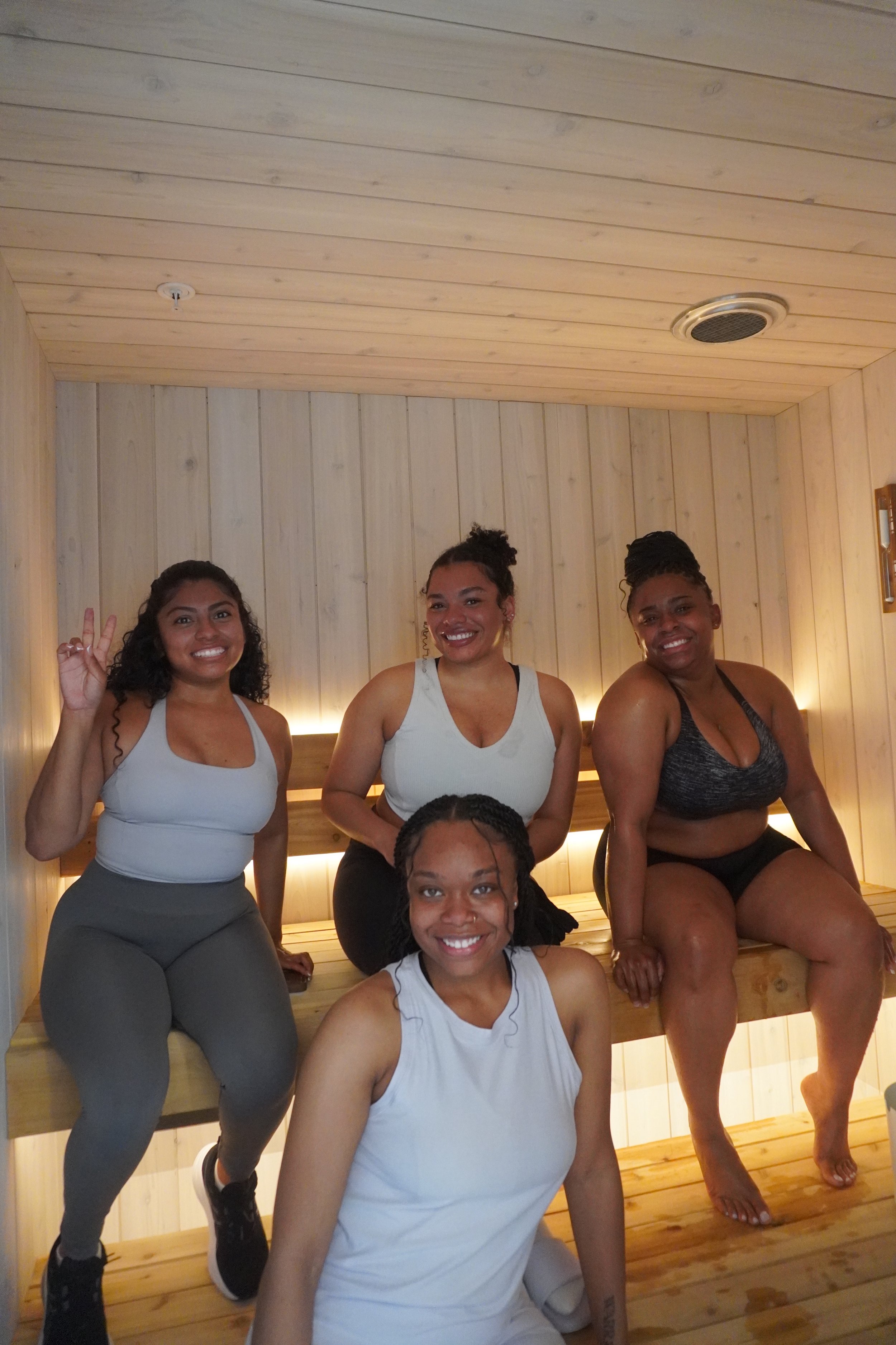 Four women sitting inside a wooden sauna, smiling at the camera, one making a peace sign.