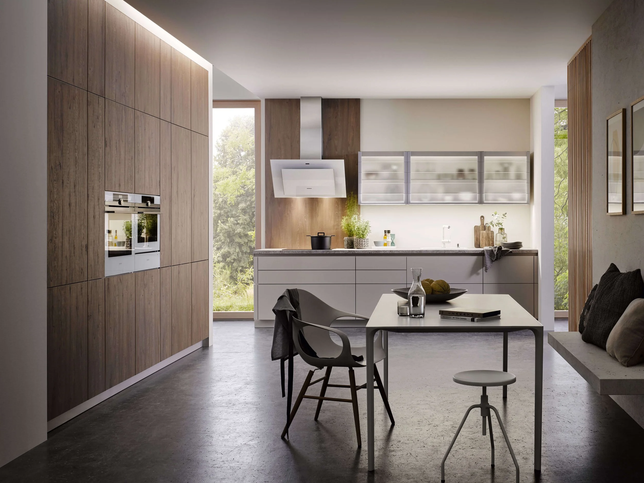 Modern kitchen with white cabinets, wooden accents, and large windows showing greenery outside, with a dining table and chairs in the foreground.