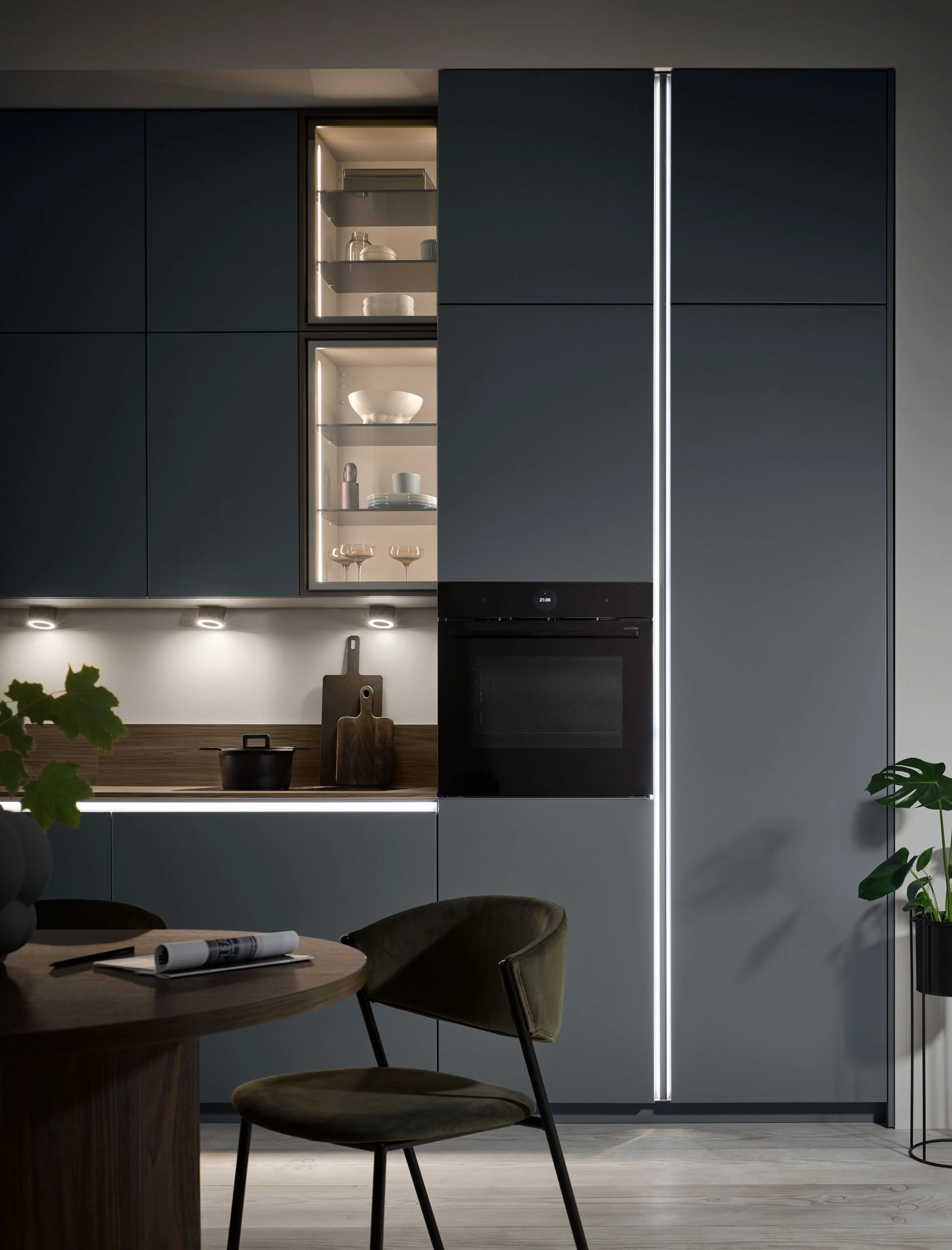Modern kitchen with dark blue cabinets, built-in oven, glass shelves with dishes, wooden countertop with cutting boards, and a dining table with green chairs.