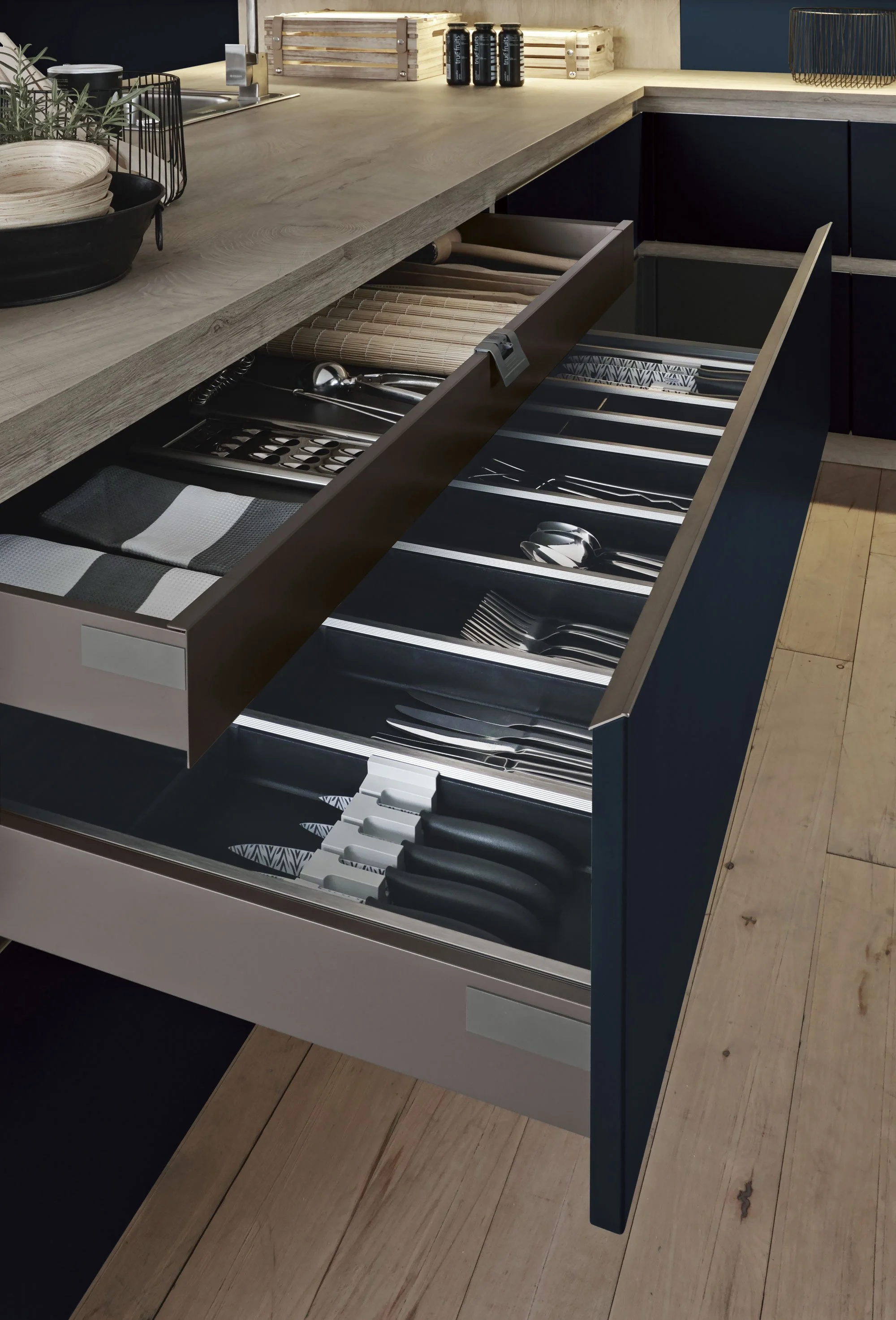Keepsake drawer filled with kitchen utensils, knives, forks, spoons, and cloth napkins, located under a wooden countertop in a modern kitchen.