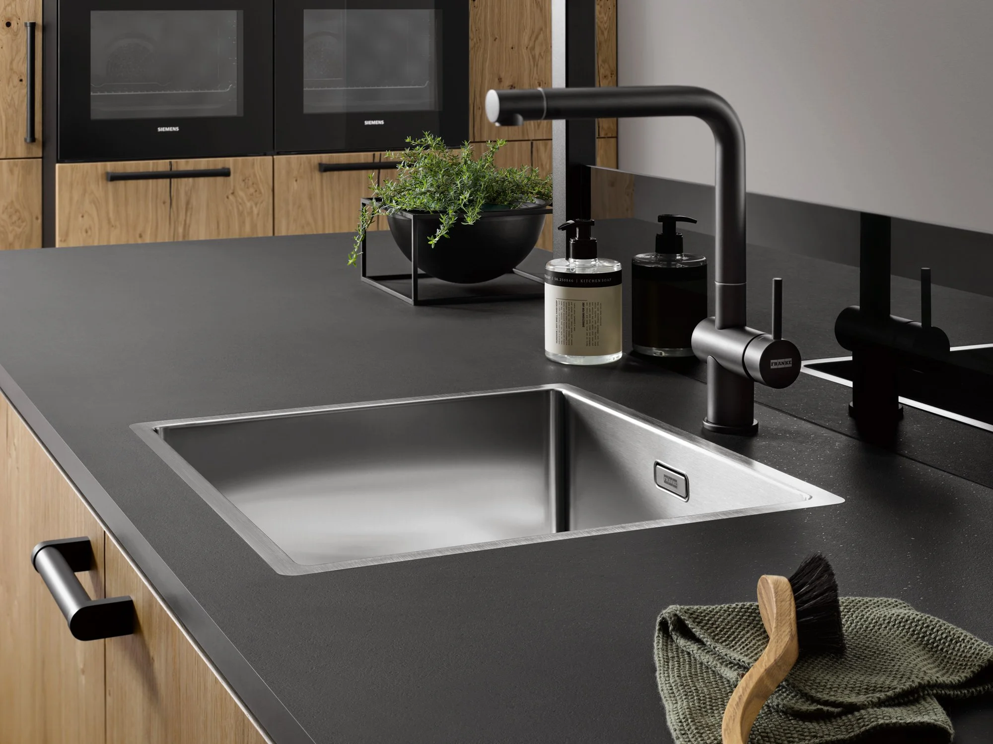 Modern kitchen sink with black tap, soap dispensers, green plant, cleaning brush, and towel on dark countertop, with wooden cabinetry and built-in ovens in the background.