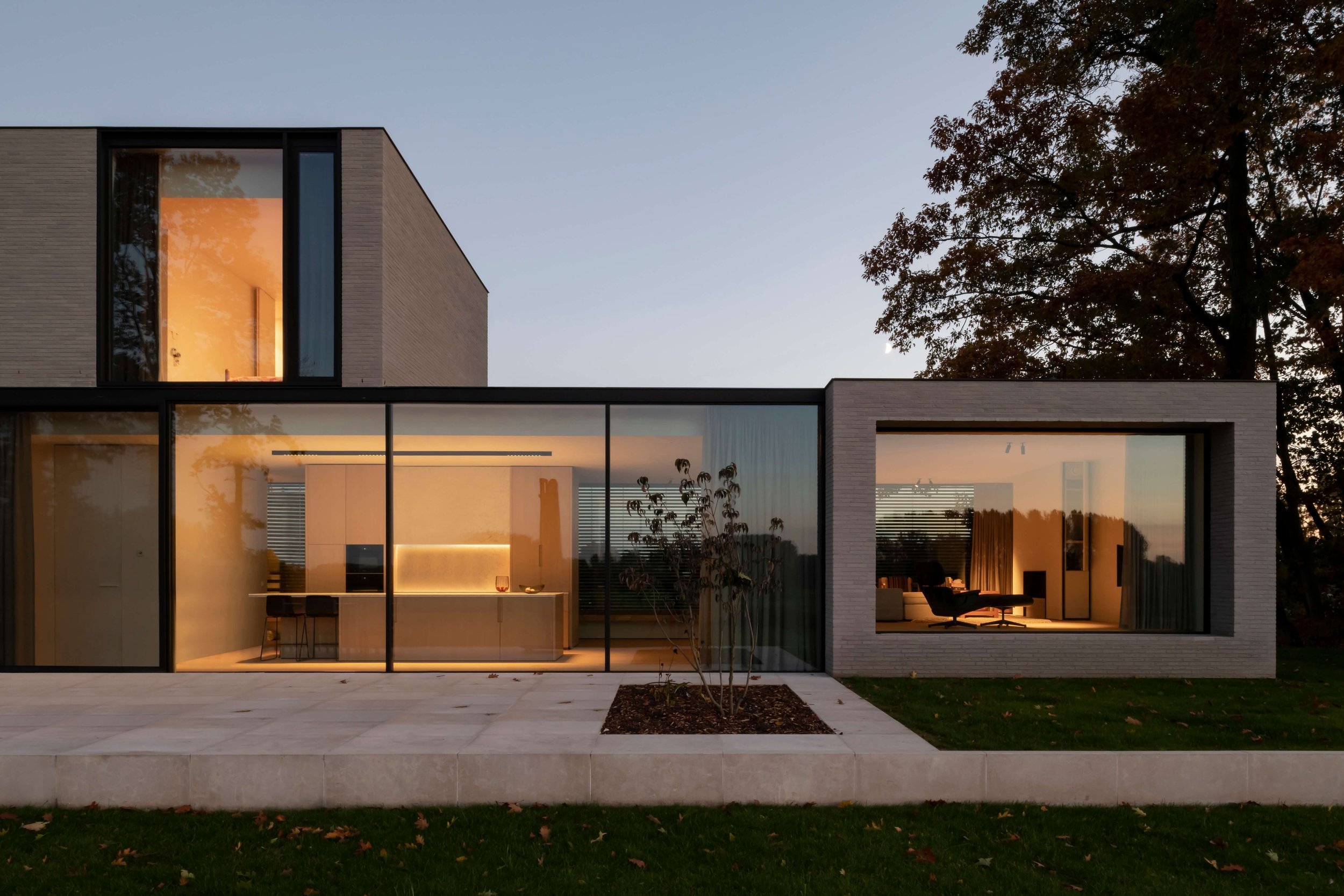Modern house with large glass windows, illuminated interior, surrounded by trees and a grassy lawn at dusk.