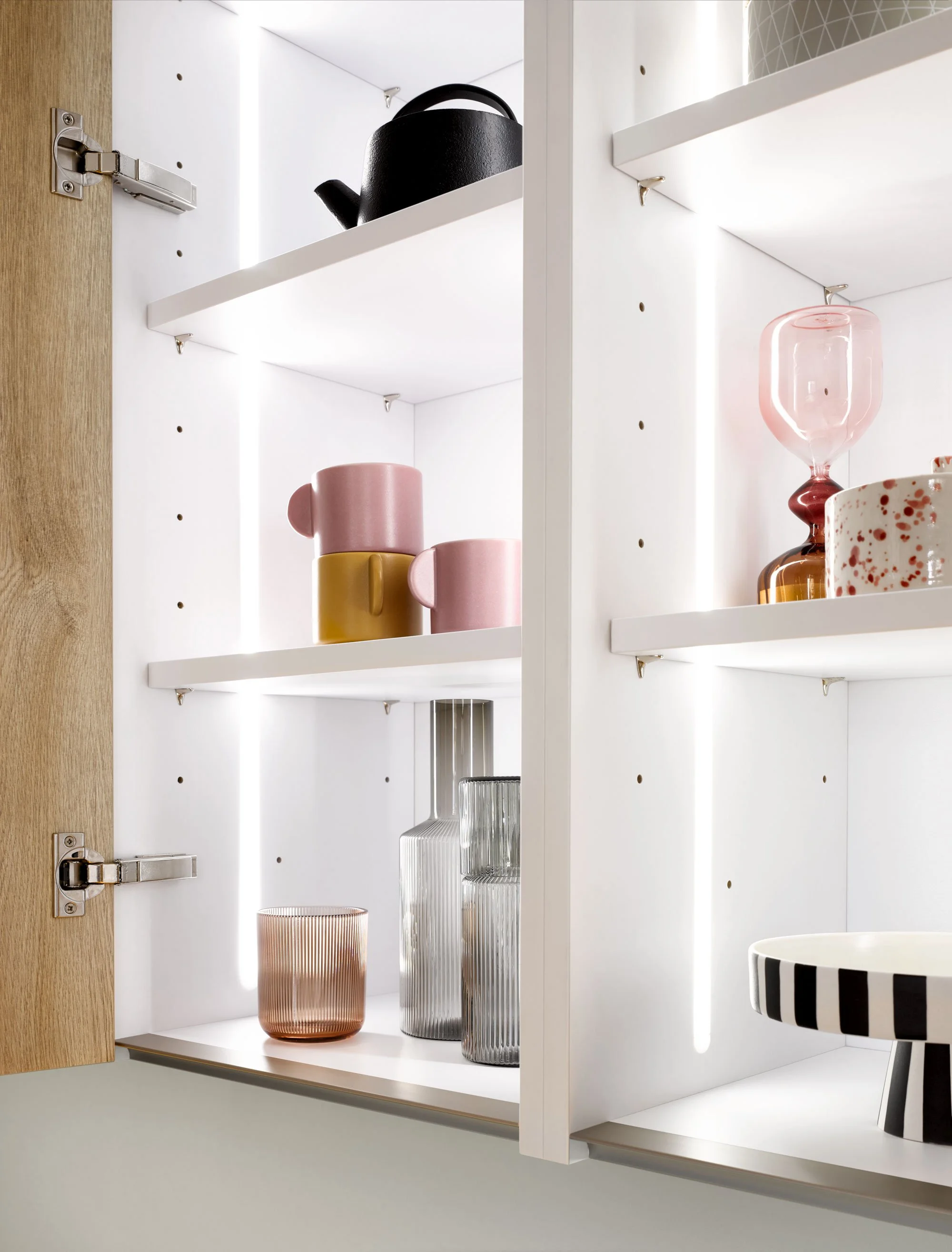 Open cabinet with glassware and mugs, including pink, black, and striped dishes, with white shelving and interior lighting.