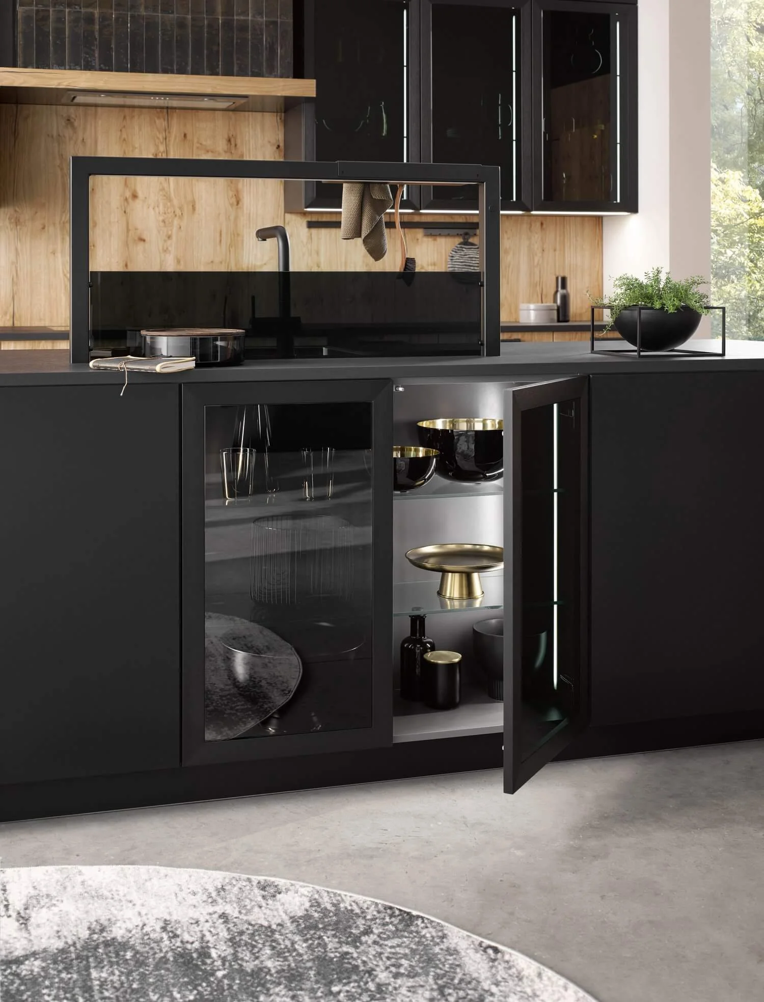 Modern kitchen with black cabinetry, wooden accents, and open display shelves showing glassware and decorative items, with a window showing greenery outside.