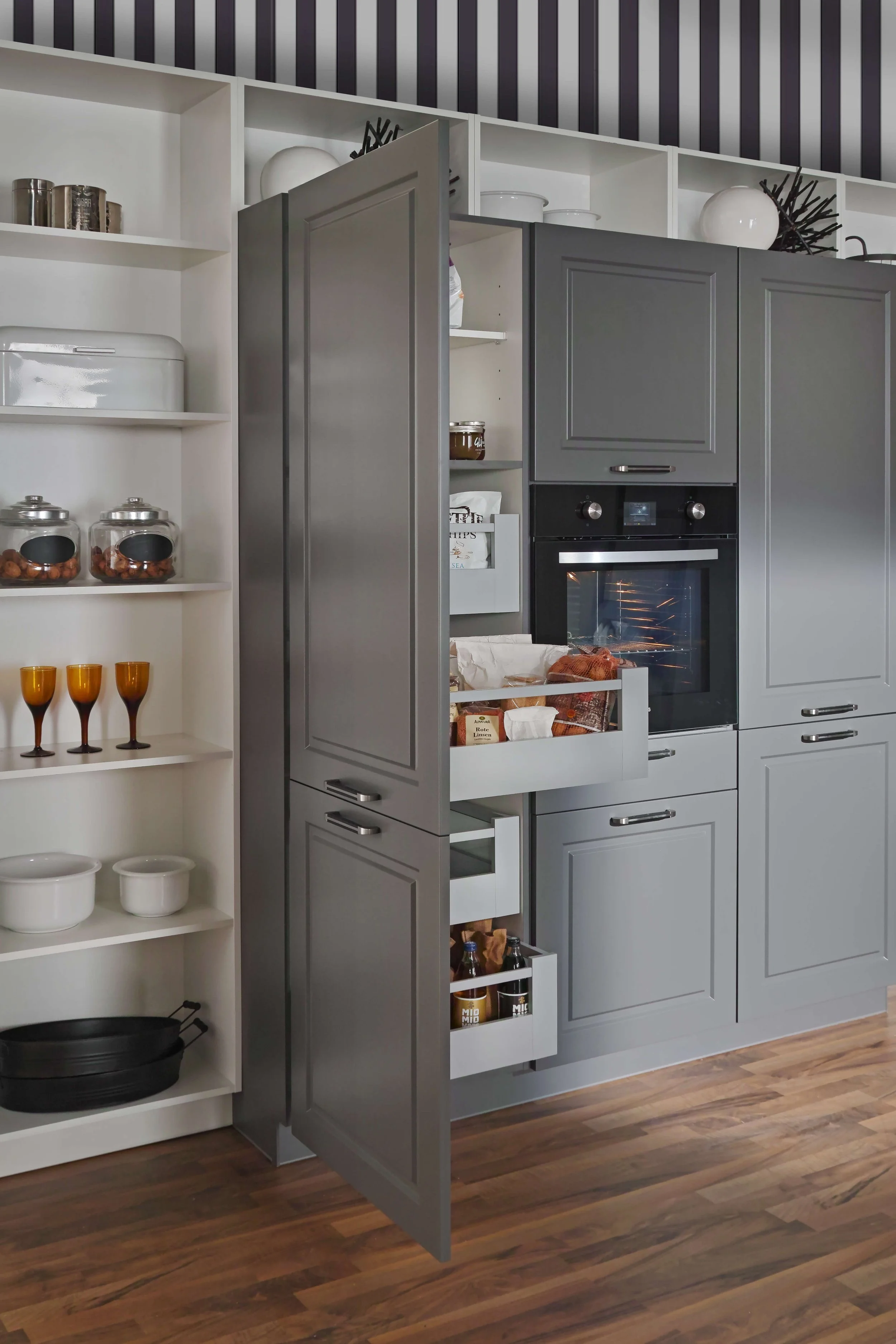 Gray kitchen cabinets with open drawers containing food items, adjacent to a white shelf with glass jars, bowls, and glasses, in a modern kitchen.