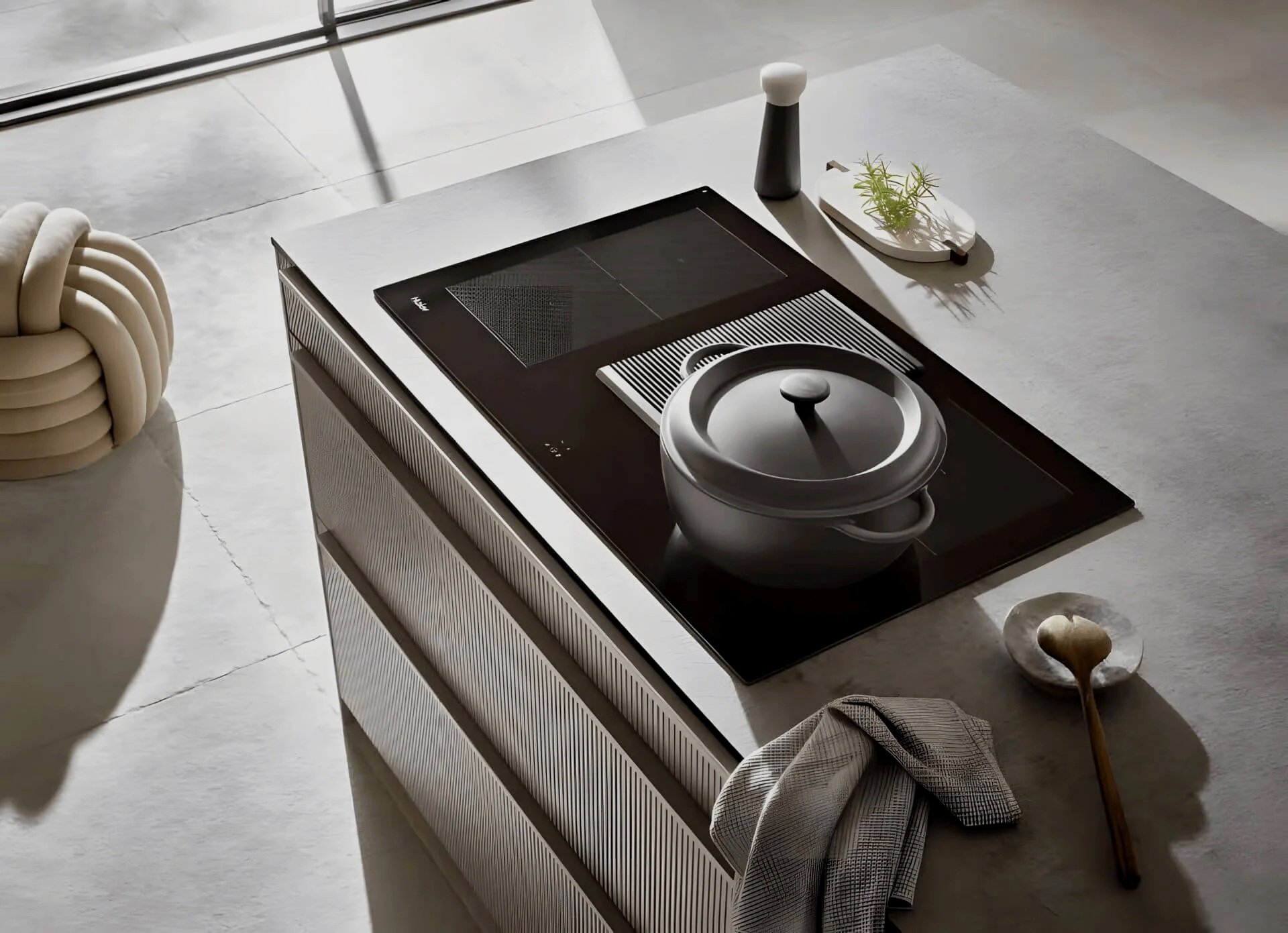 Modern kitchen countertop with a black induction cooktop, a black and white ceramic pot with a lid, a small dish with a wooden spoon, a salt shaker, and a small dish with herbs. There is also a white twisted decorative object on the side.