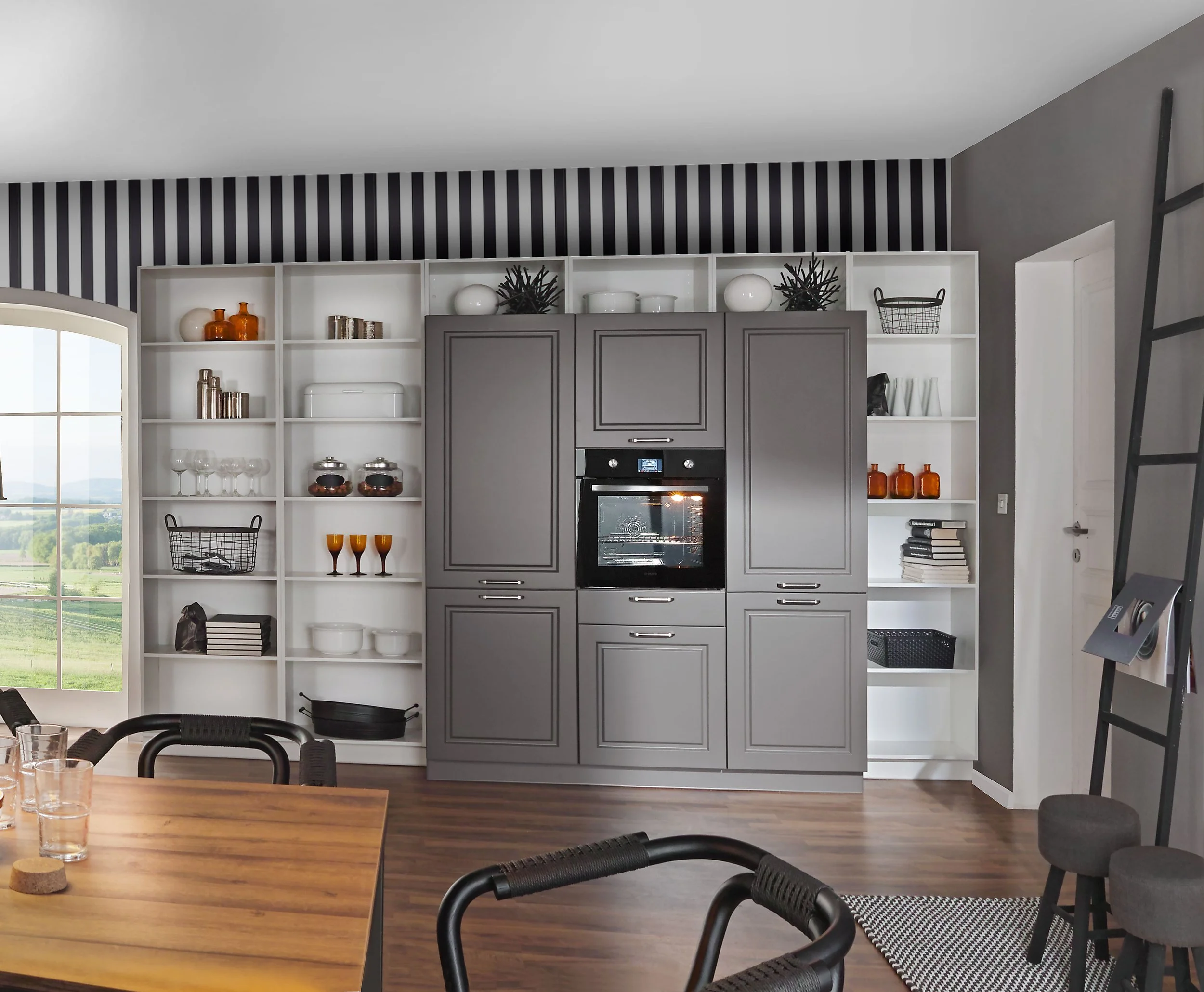 A modern kitchen with built-in gray cabinets surrounding a black oven. White open shelves on either side display glassware, decorative items, and black and amber vases. A large window on the left shows a green landscape outside, and part of a dining table with black chairs is visible in the foreground.