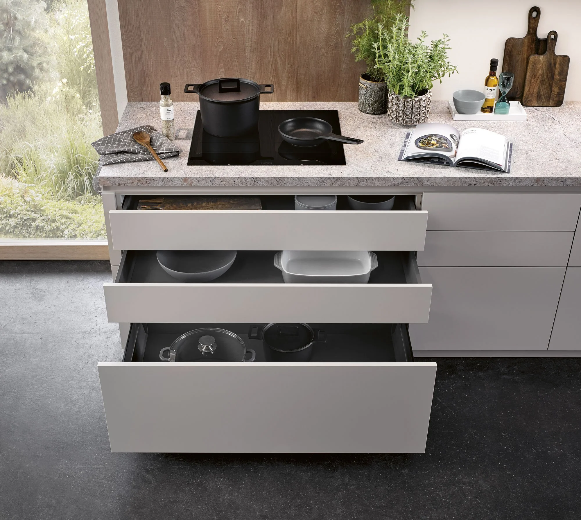 Open kitchen drawers with kitchenware, black stove with pot and pan on countertop, open cookbook, plants, cutting boards, and various condiments on a kitchen counter.