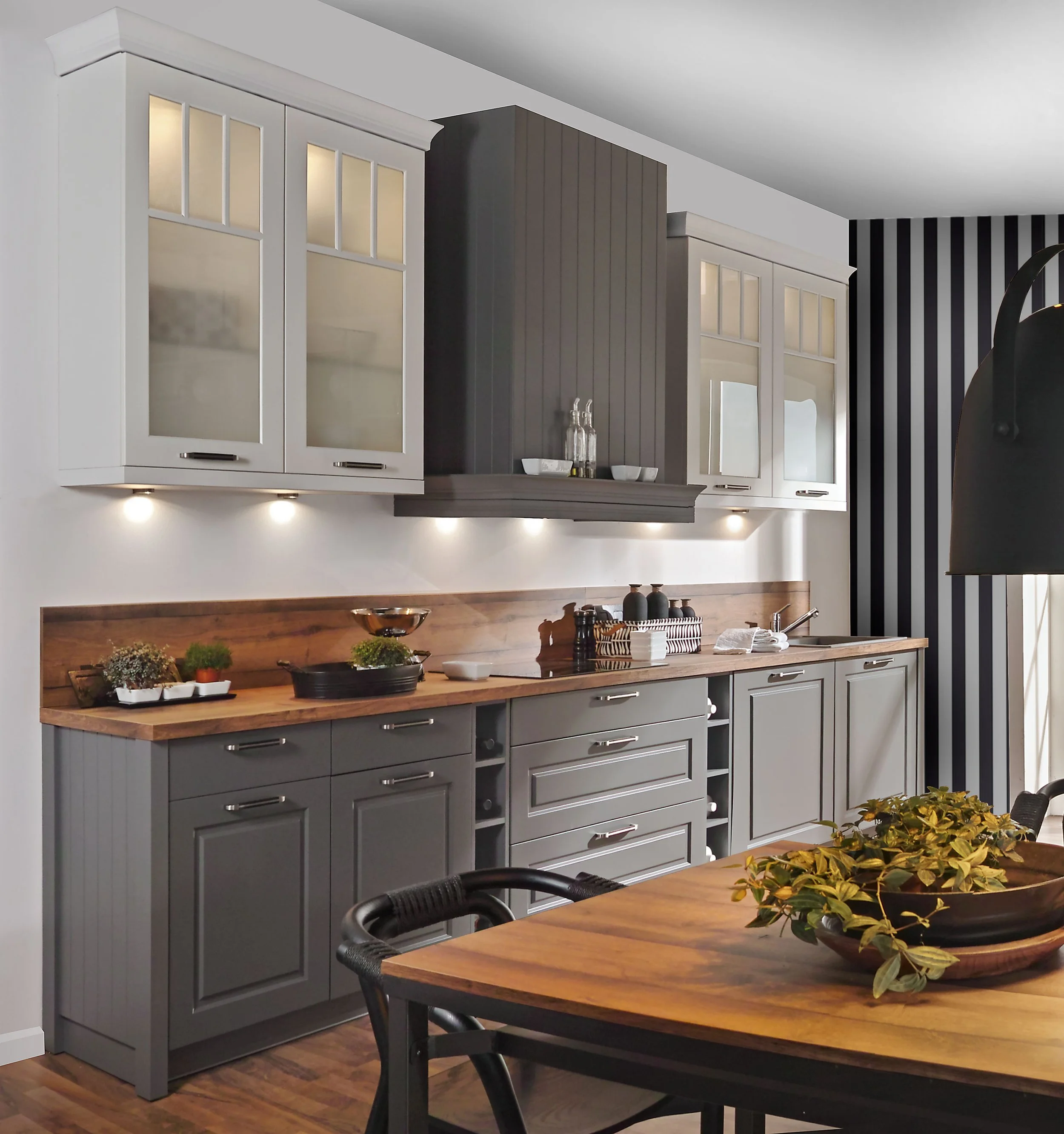Kitchen with gray lower cabinets, white upper cabinets with glass doors, wooden countertop, and a black range hood. Decor includes plants, bowls, and bottles.