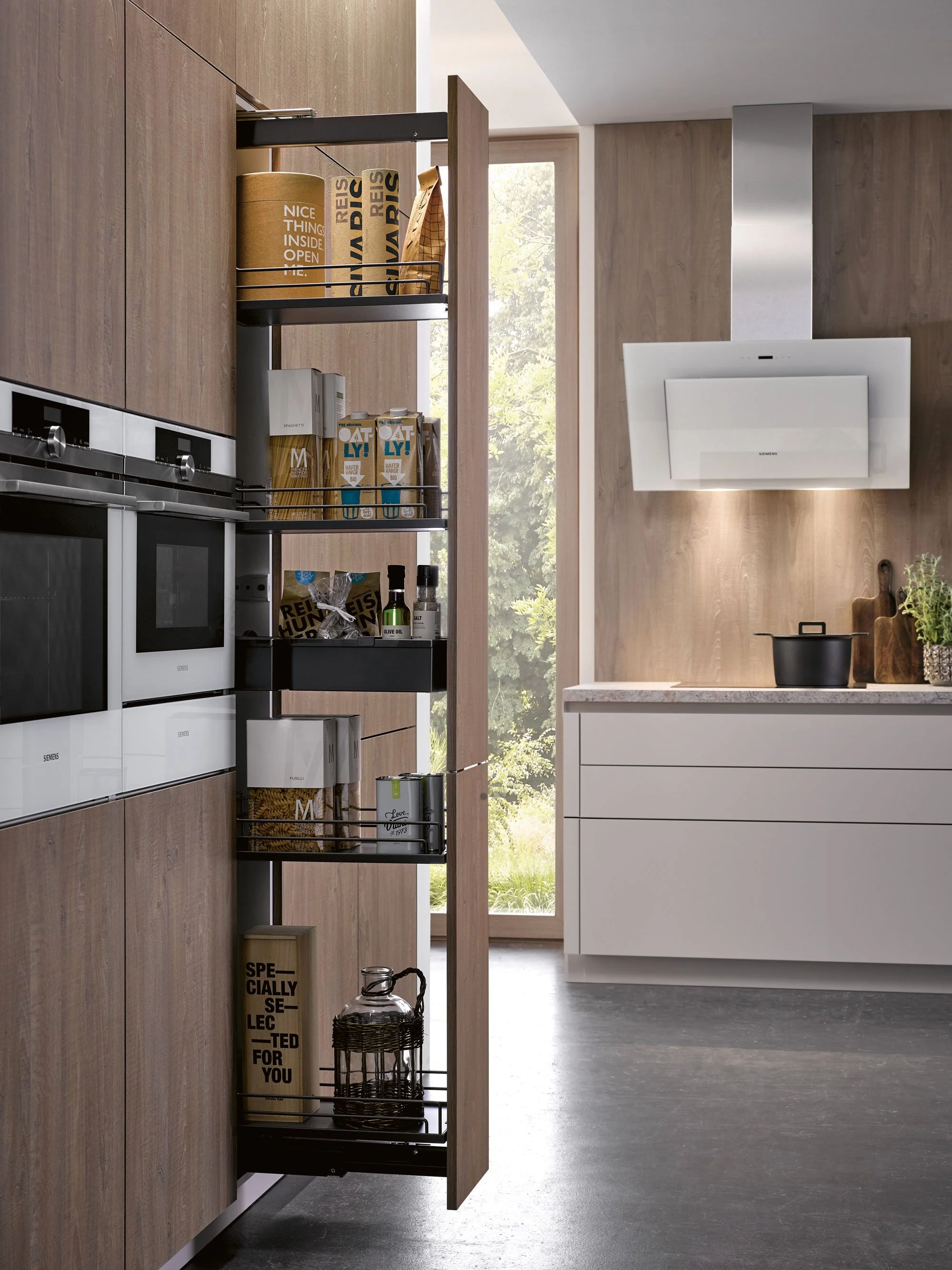 View of a modern kitchen with a wooden cabinet filled with food and kitchen items on a black metal shelf, white countertop, black pot on stove, white range hood, and large window showing greenery outside.