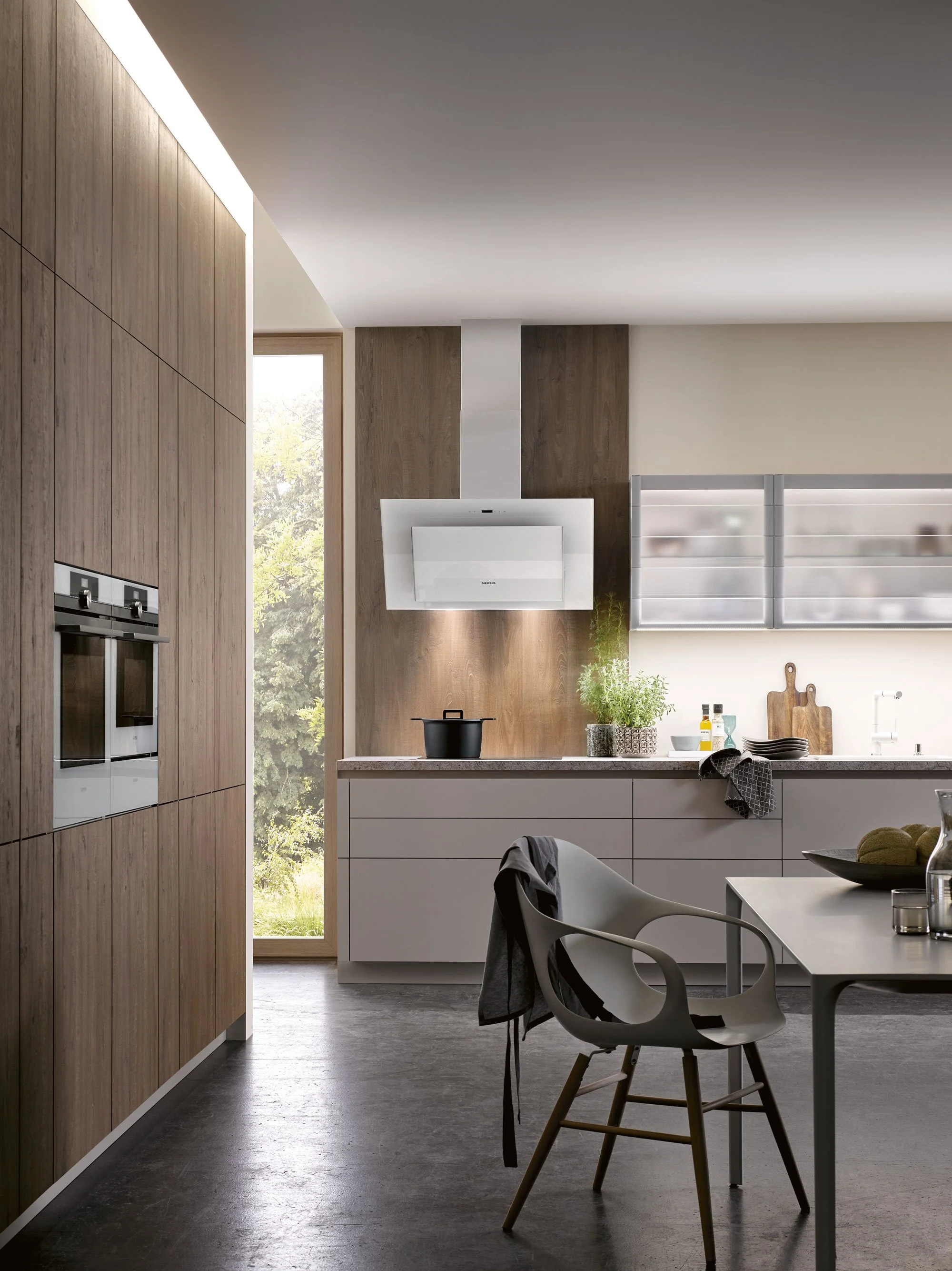 Modern kitchen with white cabinets, wooden wall panel, black cooktop, gray table, gray chair with a black cloth, window with outside greenery, potted plant, cutting boards, bottles, and bowls