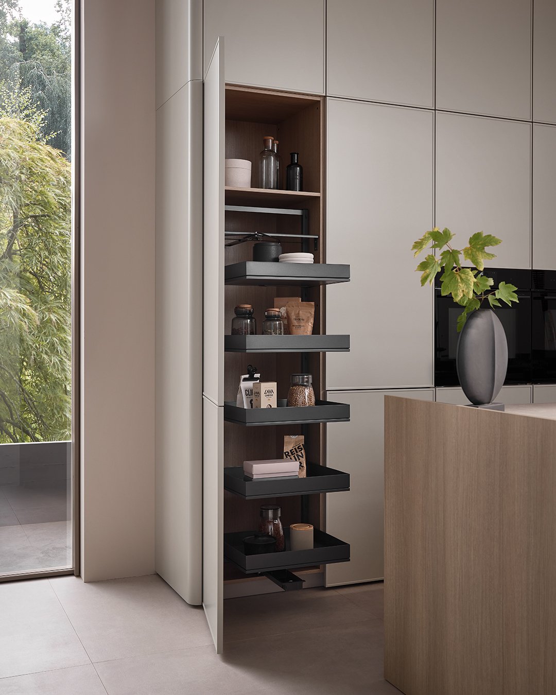Modern kitchen with a built-in pantry cabinet, open to reveal organized shelves with jars and containers, next to a window with greenery outside, and a black vase with green leaves on the counter.