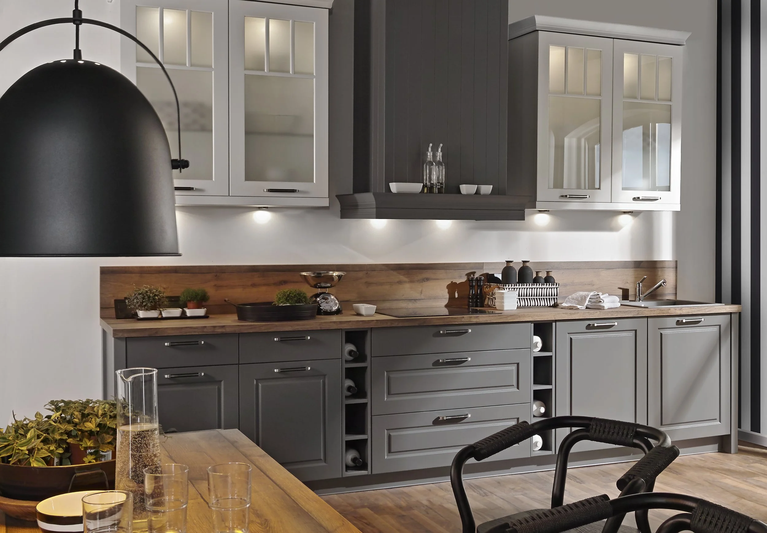 Modern kitchen with grey cabinets, wooden countertop, and white upper cabinets with glass doors. There are various kitchen items including bottles, bowls, and plants on the counter. A black pendant light hangs over a wooden dining table with glassware and a plant in the foreground.
