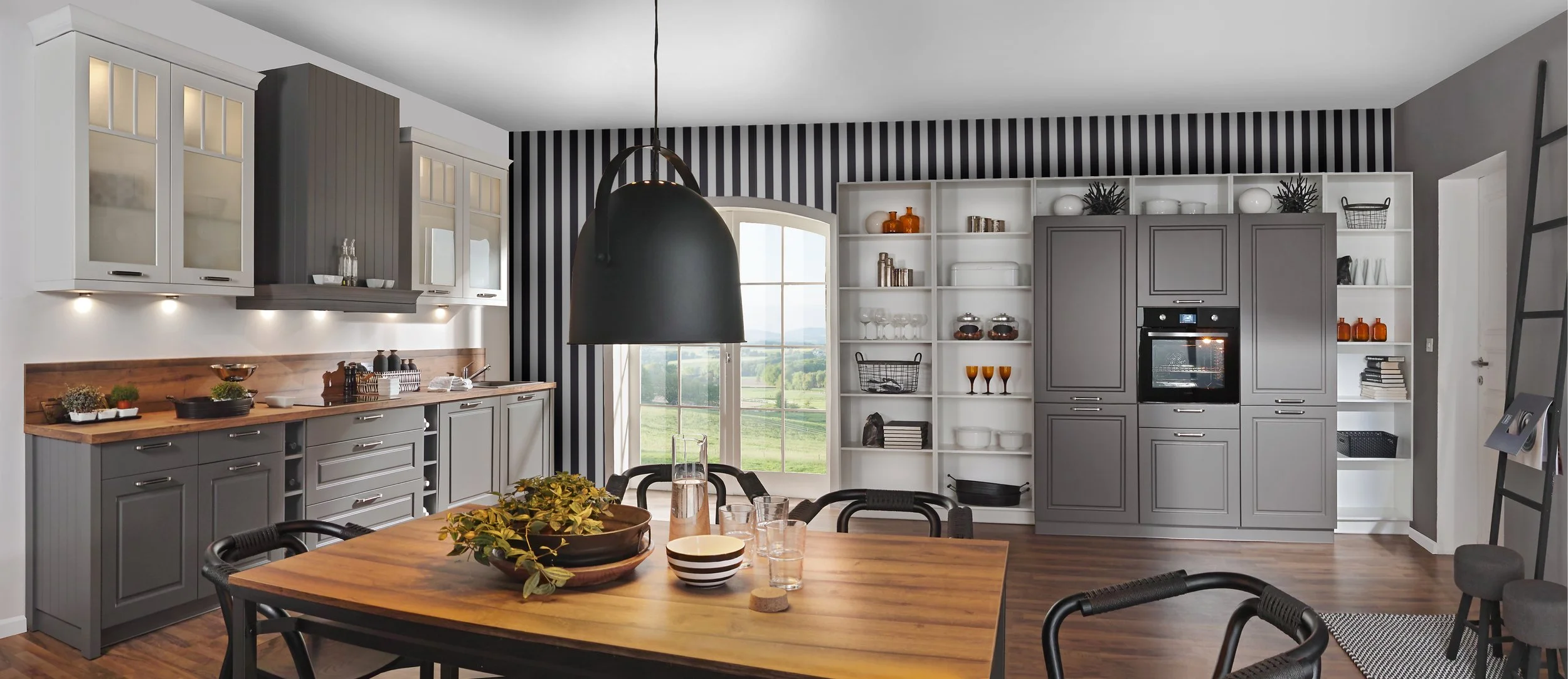 Modern kitchen with gray and white cabinetry, black and white striped wallpaper, and a large wooden dining table with black chairs. A black pendant light hangs above the table, and a window with a scenic outdoor view is in the background.