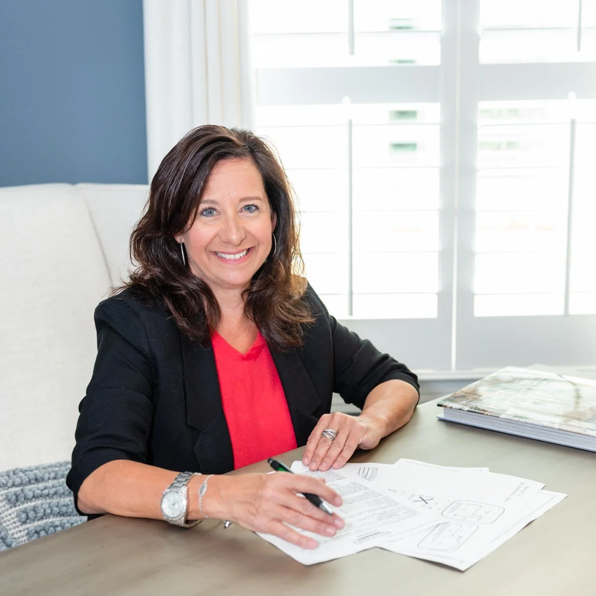 Marian Porter, Managing Partner of Next Nest Dallas, is smiling, wearing a black blazer and red top, sitting at a desk with papers and a pen, in a bright room with white shutters.