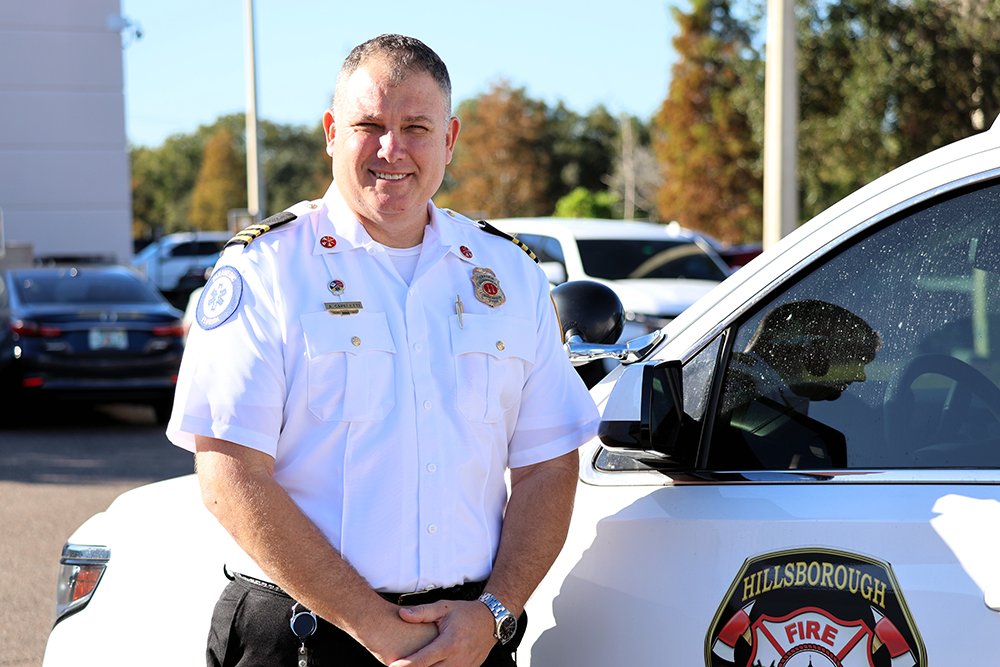 A man in a white Hillsborough Fire Department uniform standing beside a white vehicle with a fire department badge on the door.