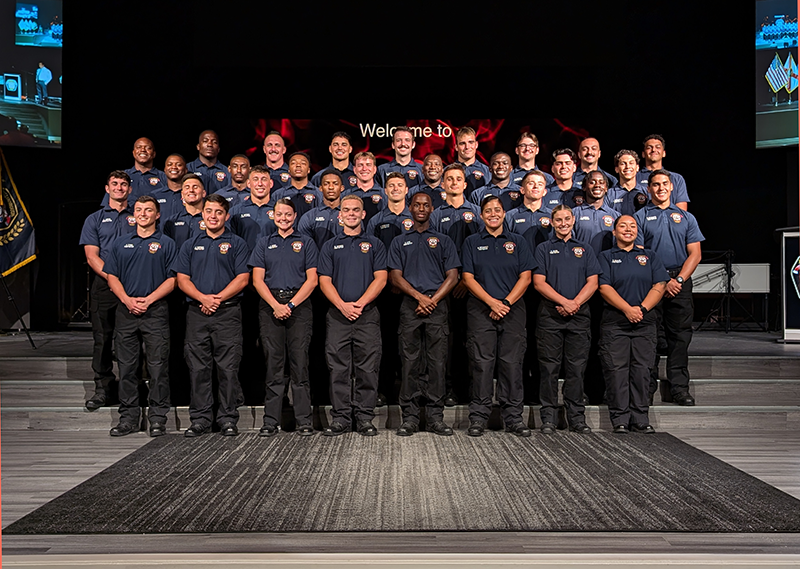 Group of uniformed individuals posing on stage at a formal event, with a red and black background and a sign that reads 'Welcome to'.