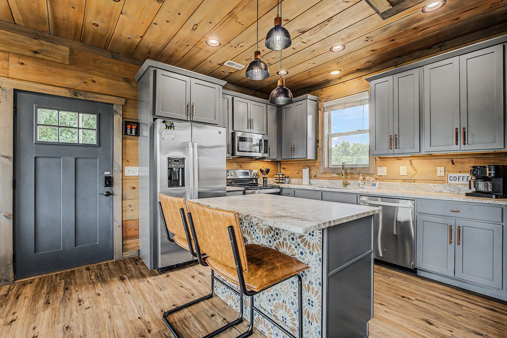 A modern kitchen with gray cabinets, stainless steel refrigerator, microwave, and stove, wood-paneled ceiling and walls, pendant lights, a window, and a countertop breakfast bar with wooden chairs.
