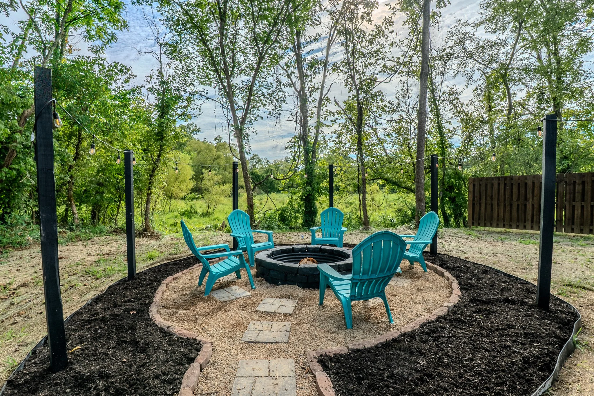 Outdoor backyard fire pit area with six turquoise Adirondack chairs arranged in a circle around a fire pit, with string lights overhead and surrounded by trees and greenery.