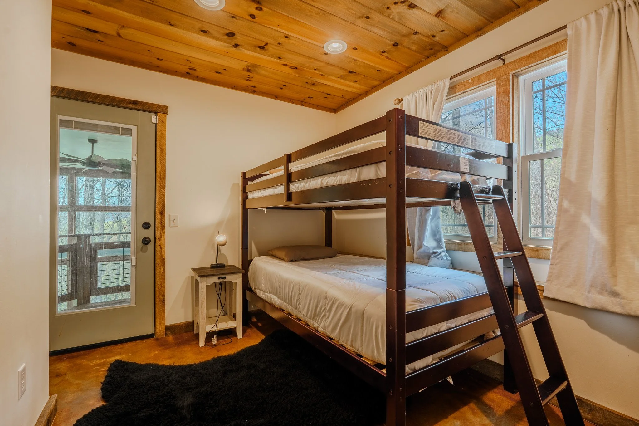 A bedroom with a bunk bed positioned near windows with white curtains, a side table with a lamp, and a door leading outside. The ceiling is made of wood planks, and the floor has a black rug.