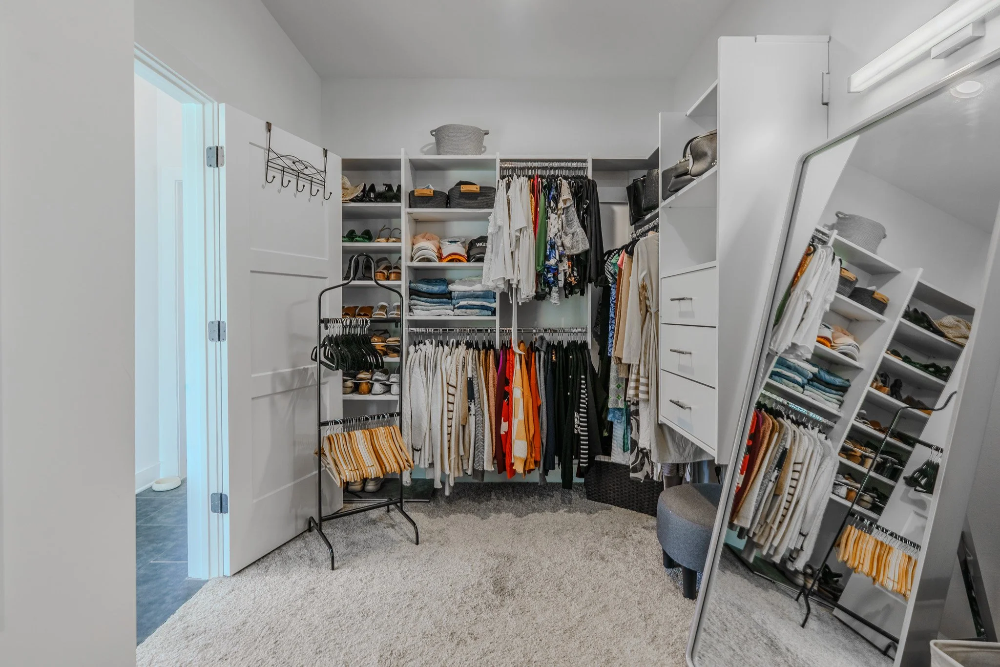 A walk-in closet with white shelving units filled with folded clothes, shoes, bags, and hanging garments. A large mirror leans against the right wall, reflecting part of the closet and a gray stool. The floor is covered with light beige carpet.