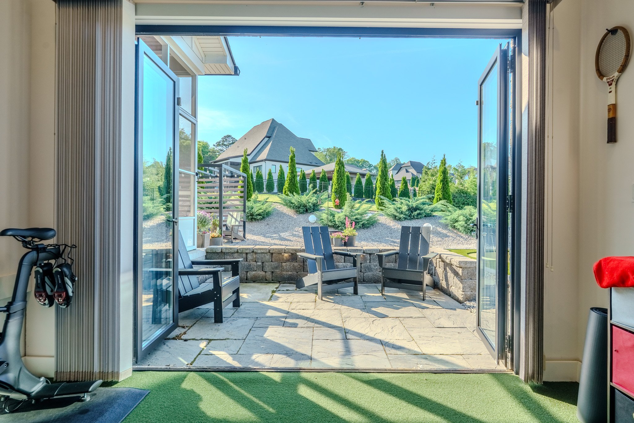 Open patio door leading to backyard with black chairs, potted plants, and landscaping with green bushes and trees, and neighboring houses under a clear blue sky.