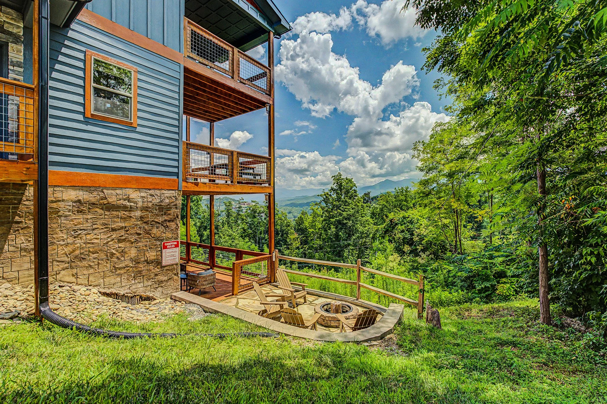 A multi-story house with blue siding, wooden balconies, and stone foundation, surrounded by trees and greenery on a hill under a partly cloudy sky.