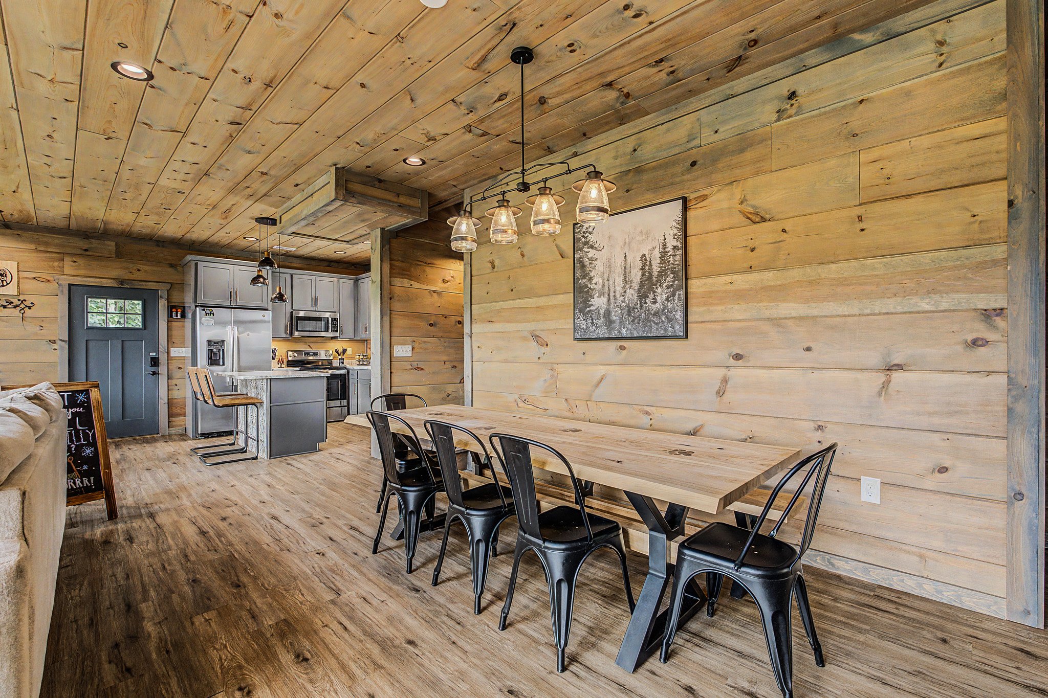 Open-concept kitchen and dining area with wooden walls and ceiling, black dining chairs, a large wooden dining table, and modern kitchen appliances including a microwave and refrigerator.