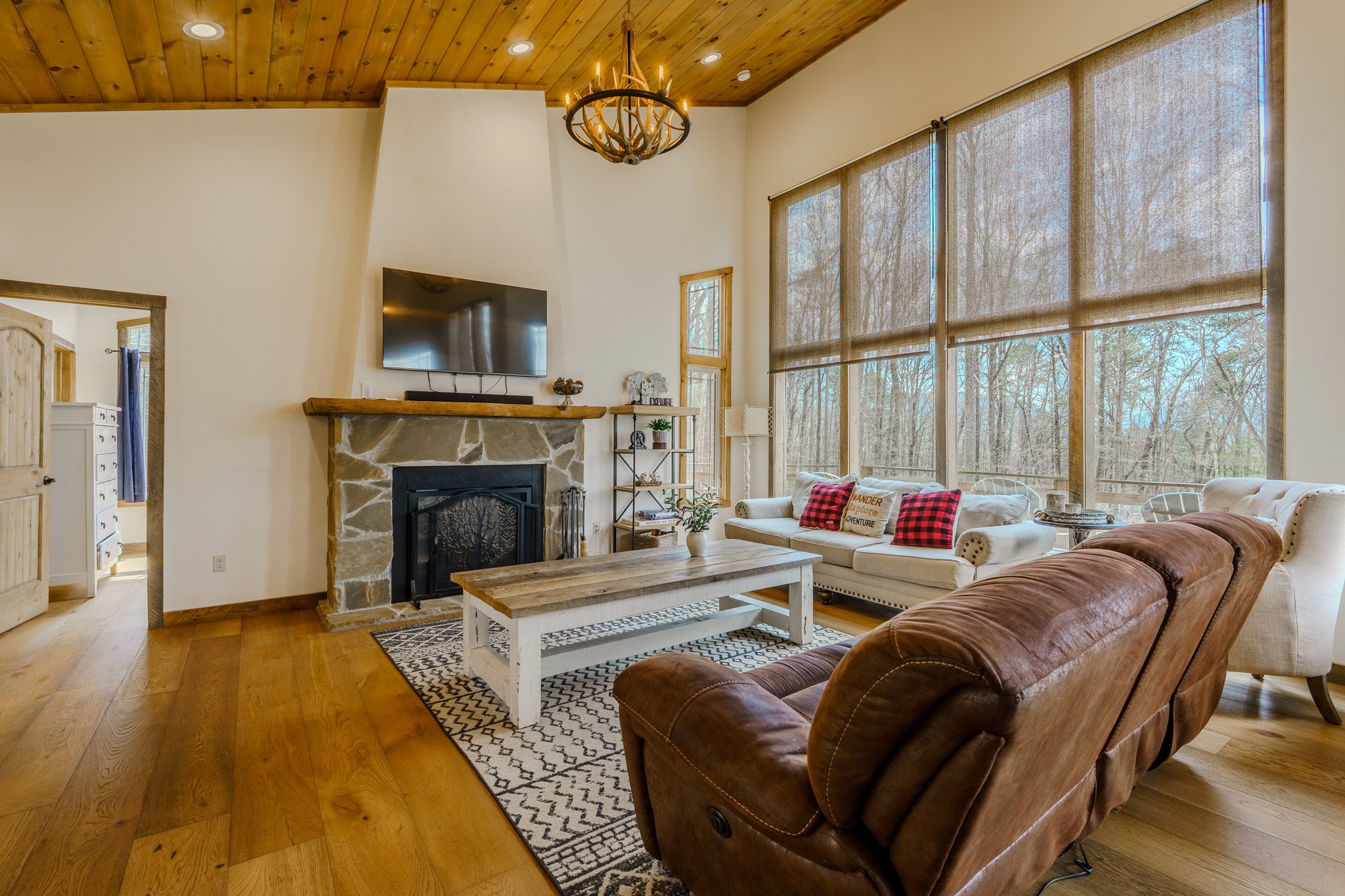 Living room with a leather sofa, white sofa, wooden coffee table, stone fireplace, flat-screen TV, large windows with blinds, wooden ceiling, and decorative chandelier.
