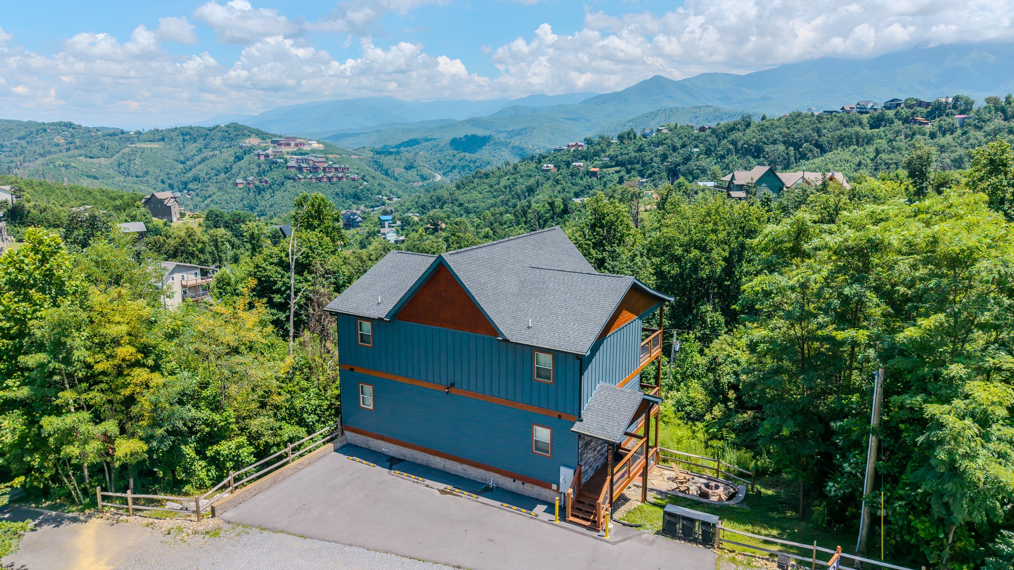 A house with blue siding and a gray roof situated on a hillside surrounded by green trees, with mountainous terrain and a partly cloudy sky in the background.