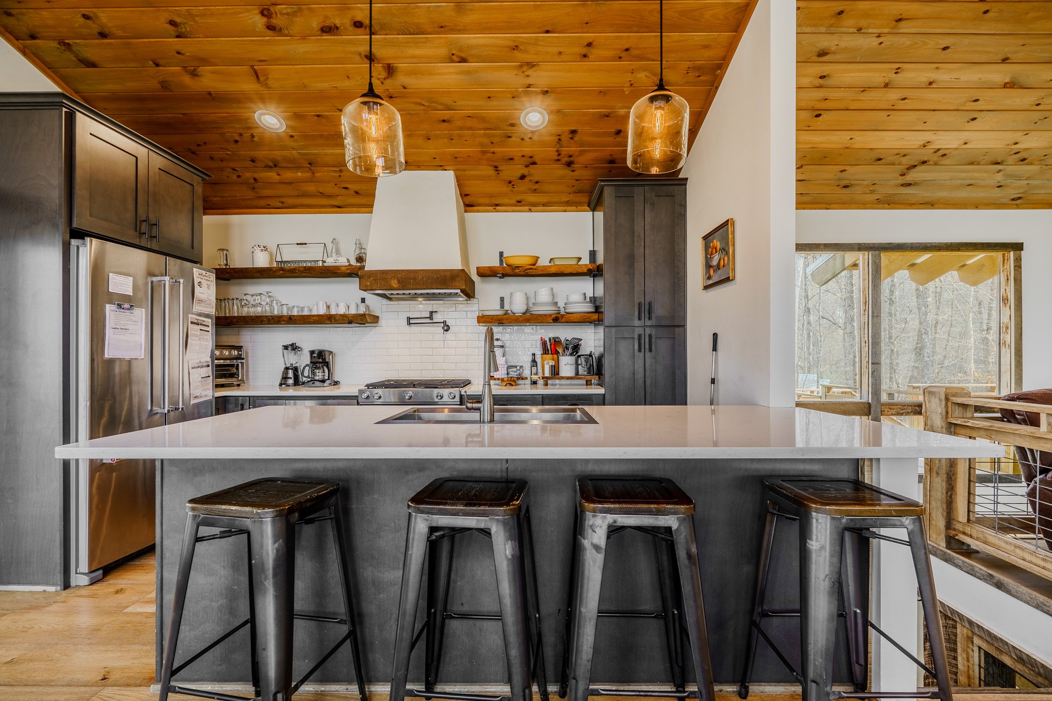 Modern kitchen with wooden ceiling, black cabinets, open shelves with dishes, stainless steel fridge, and a white island with four metal bar stools. Large windows showing outdoor wooden deck and trees.
