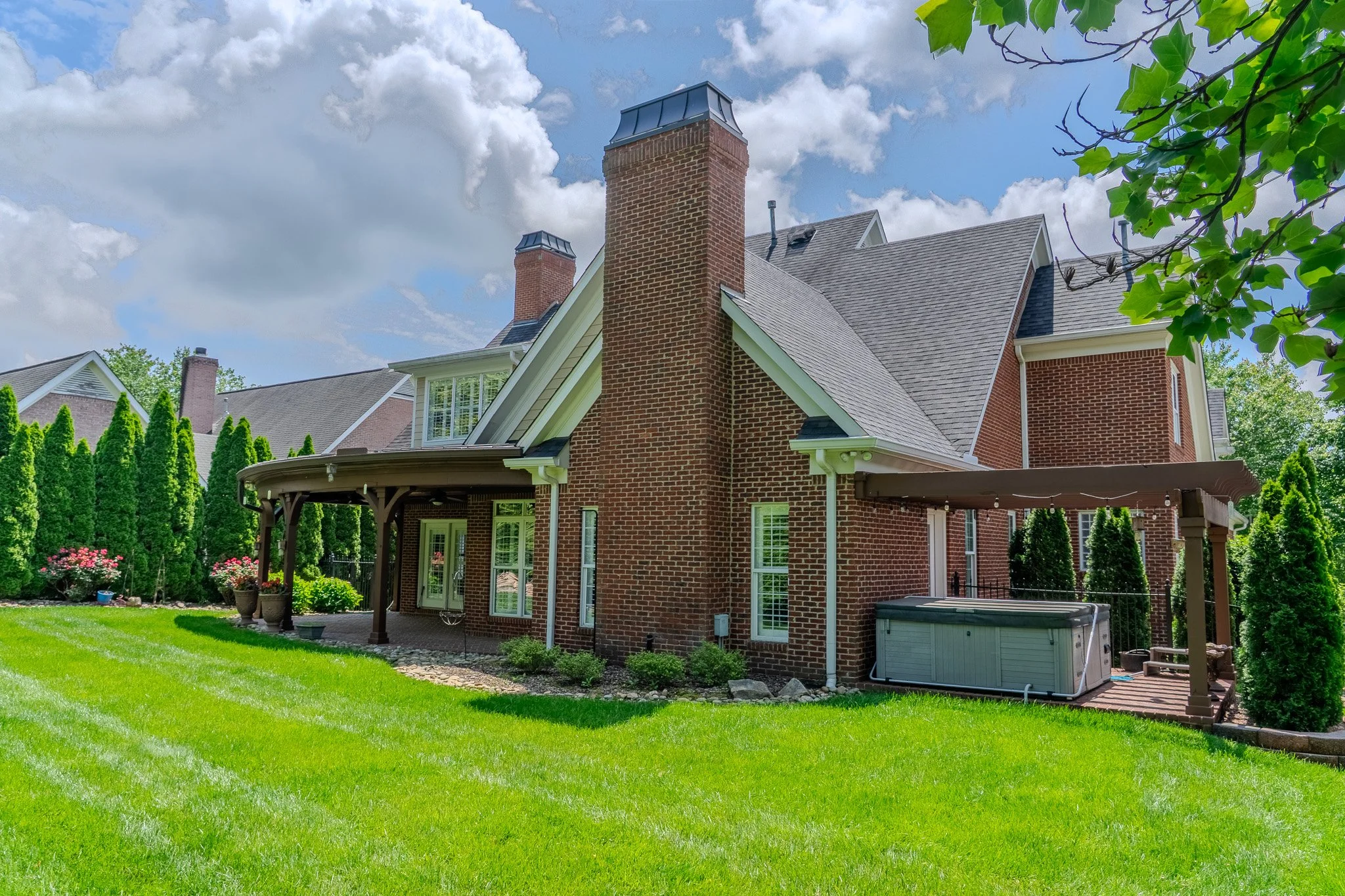 Rear view of a brick house with a chimney, covered patio, and a garden hot tub on a wooden deck, surrounded by a lawn and tall evergreen trees.