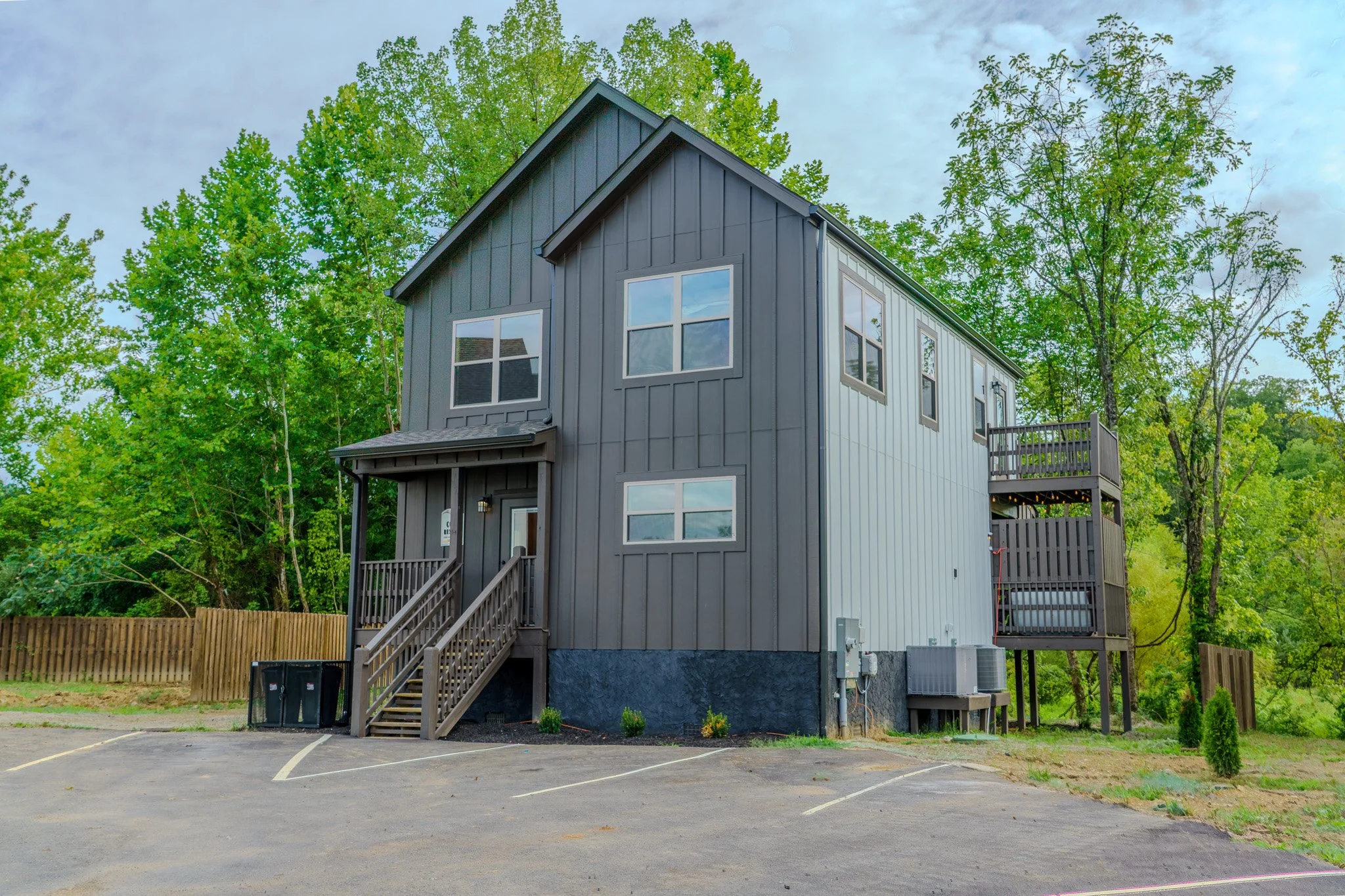 front view of a modern, two-story house with black and white siding, surrounded by green trees, with a parking lot in the foreground.