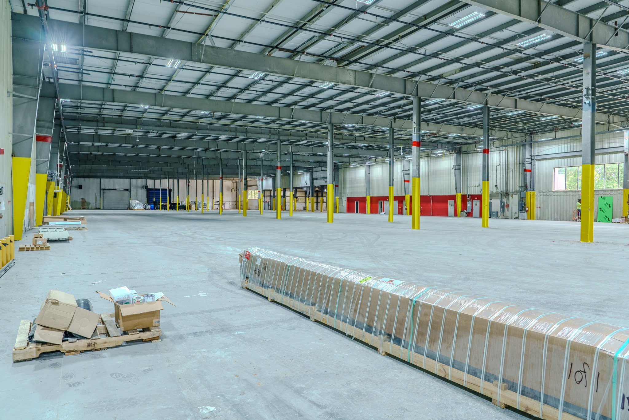 Interior of a large, empty warehouse under construction with yellow safety poles, stacks of packaging materials, and a partially installed ceiling.