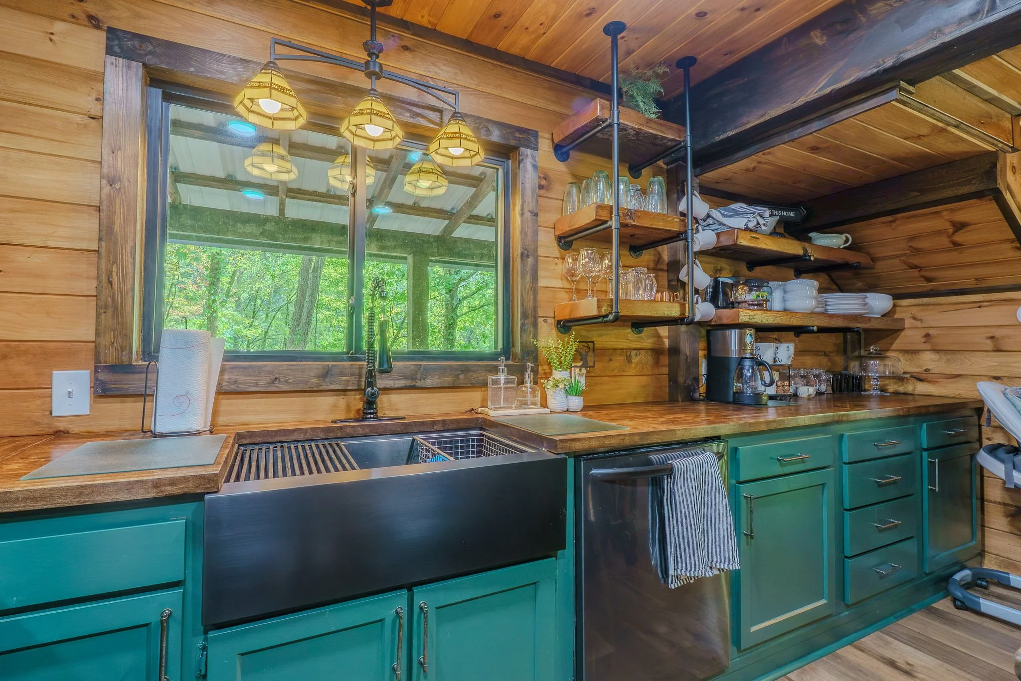 Kitchen with wooden walls, teal cabinets, open wooden shelves with dishes, glasses, and mugs, a window with a view of green trees, and a sink with a window above it.