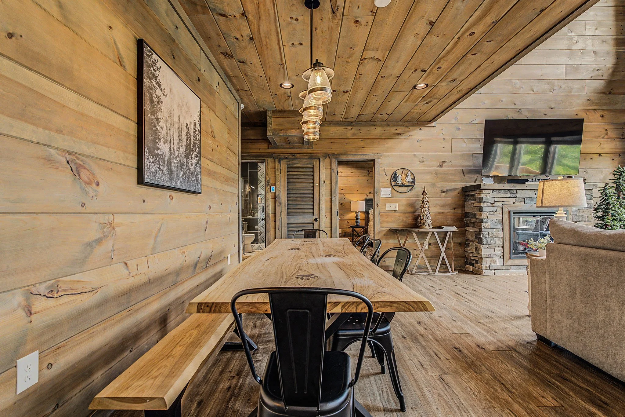 Interior of a rustic wooden room with a long wooden dining table, black chairs, a stone fireplace, a mounted flat-screen TV, and decorative items including a small Christmas tree and holiday-themed decor.