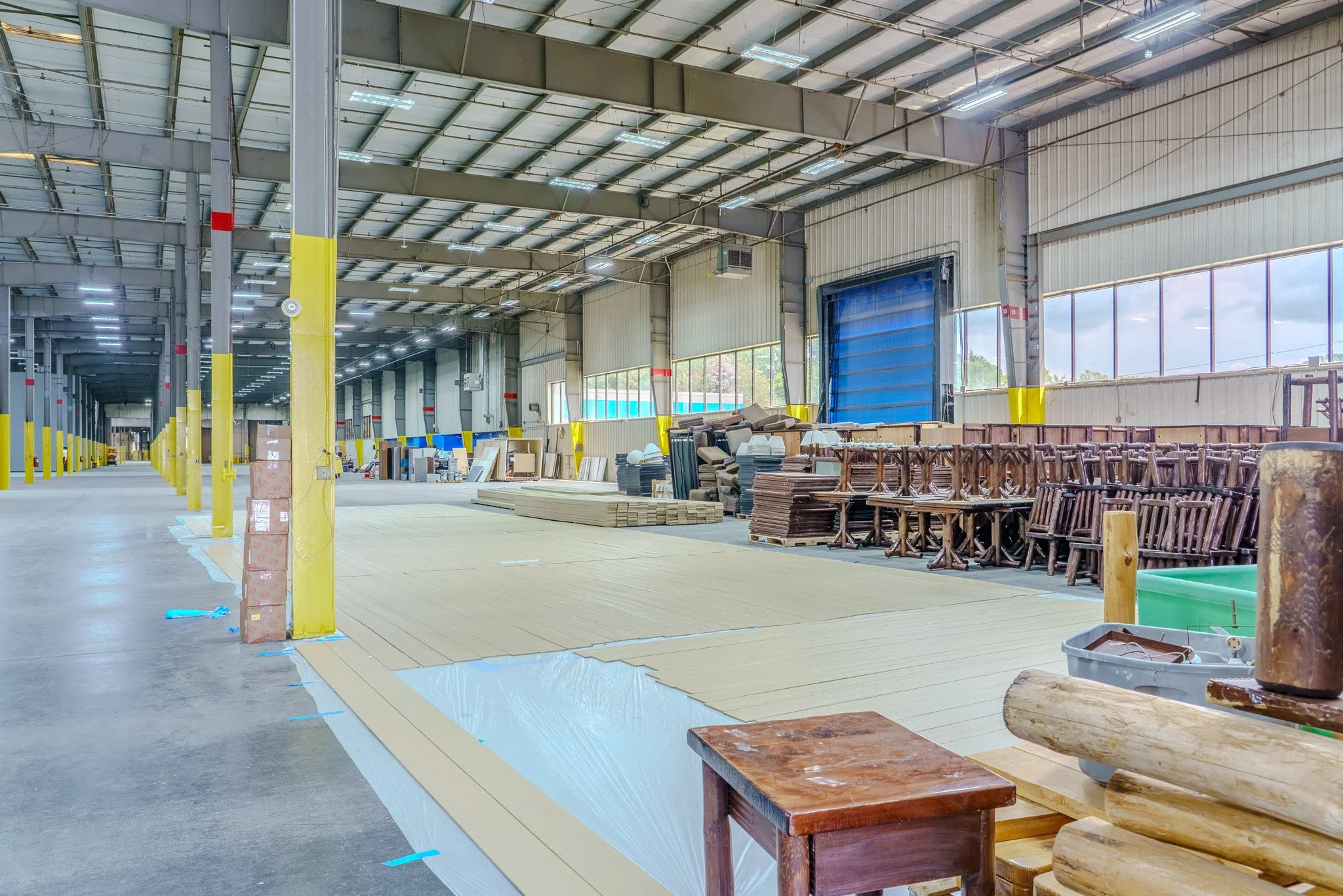 Interior view of a warehouse under renovation with stacks of wooden chairs, furniture pieces, and construction materials, featuring high ceilings and large windows.