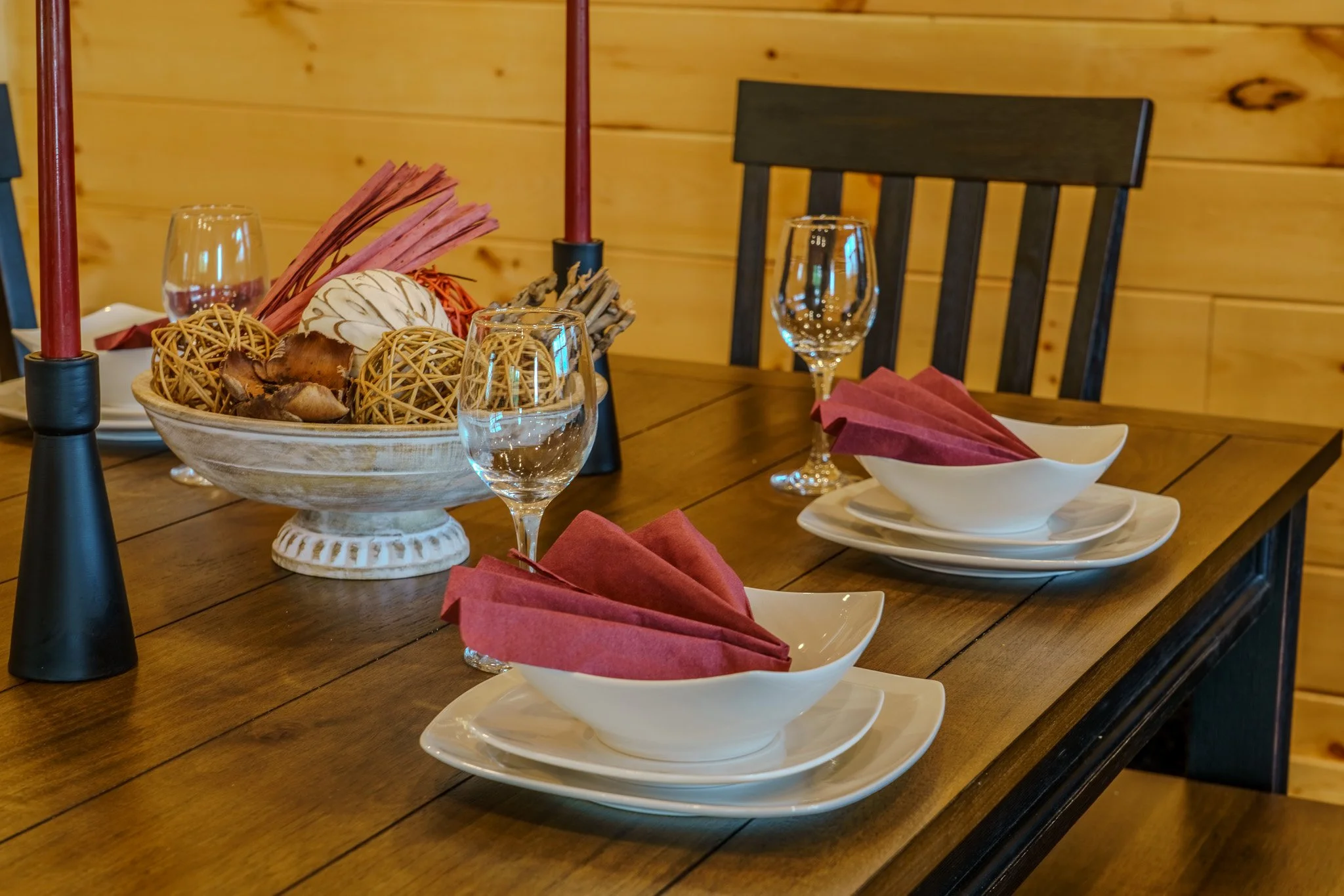 A wooden dining table set for a meal, with white plates and wine glasses, pink napkins in white bowls, decorative candles, and a centerpiece with orbs and dried foliage, against a wood-paneled wall.