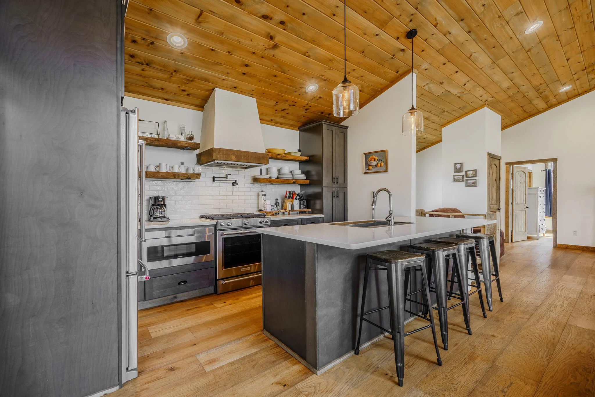 Modern kitchen with wooden ceiling, white walls, stainless steel appliances, black kitchen island, open shelves with dishes, and wooden flooring.
