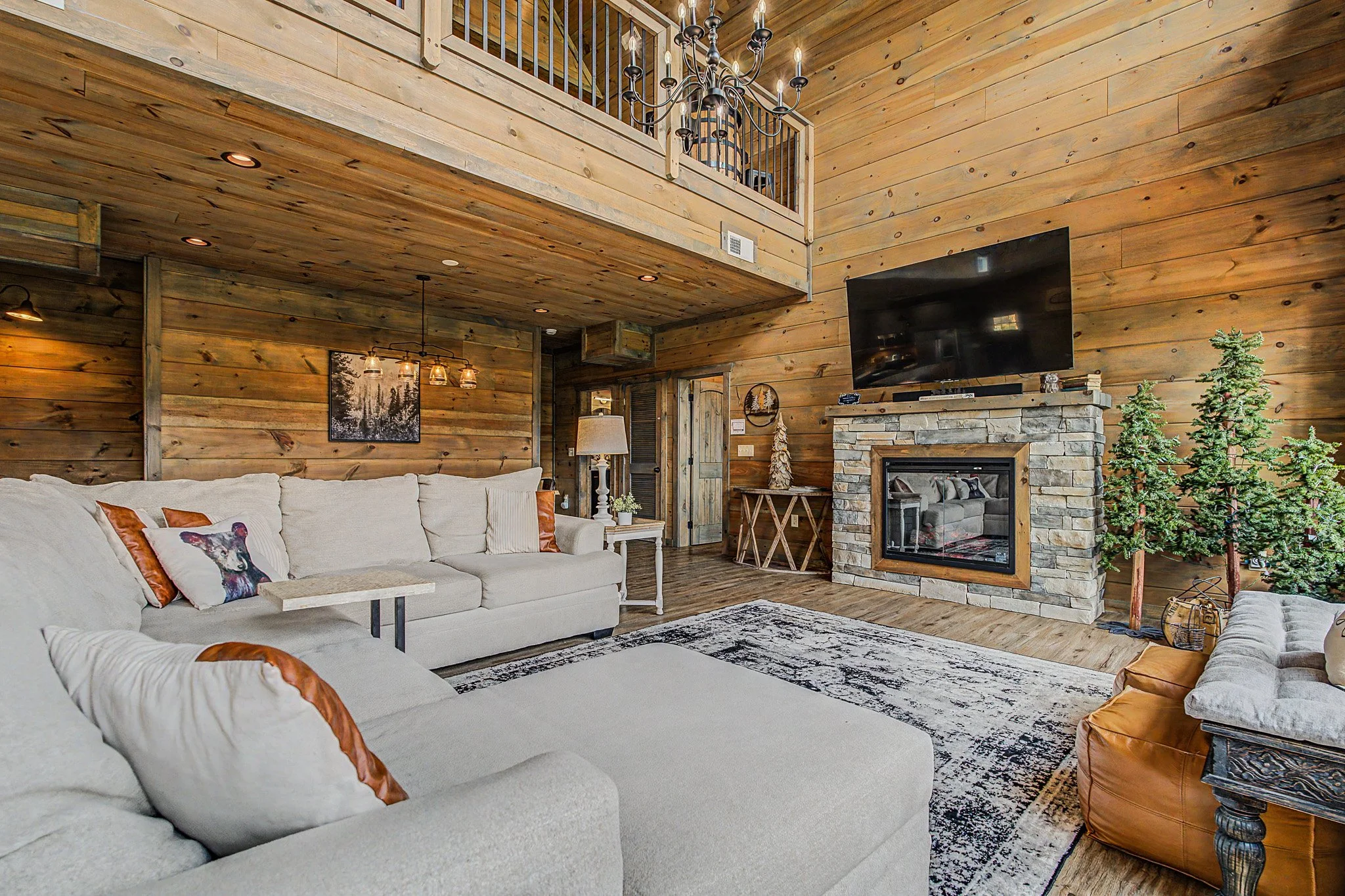 Interior of a cozy living room with wooden walls, a fireplace, and a large flat-screen TV mounted above it. There is a beige sofa with decorative pillows featuring animal prints, including a dog. The room is decorated with small trees, a rug, and war