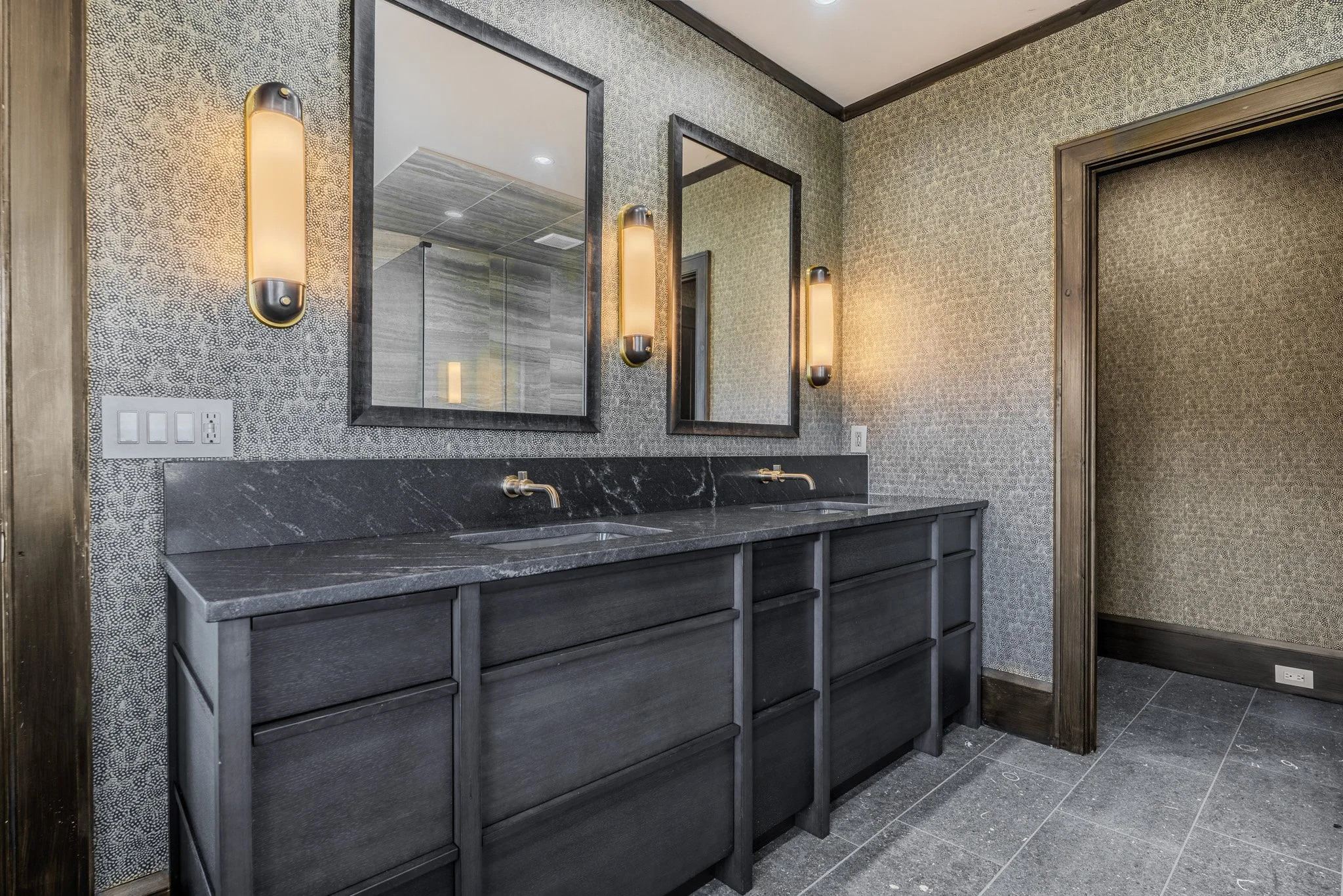Double bathroom vanity with black countertops, two sinks, two mirrors, and modern wall sconces, in a bathroom with textured wallpaper and dark wooden accents.