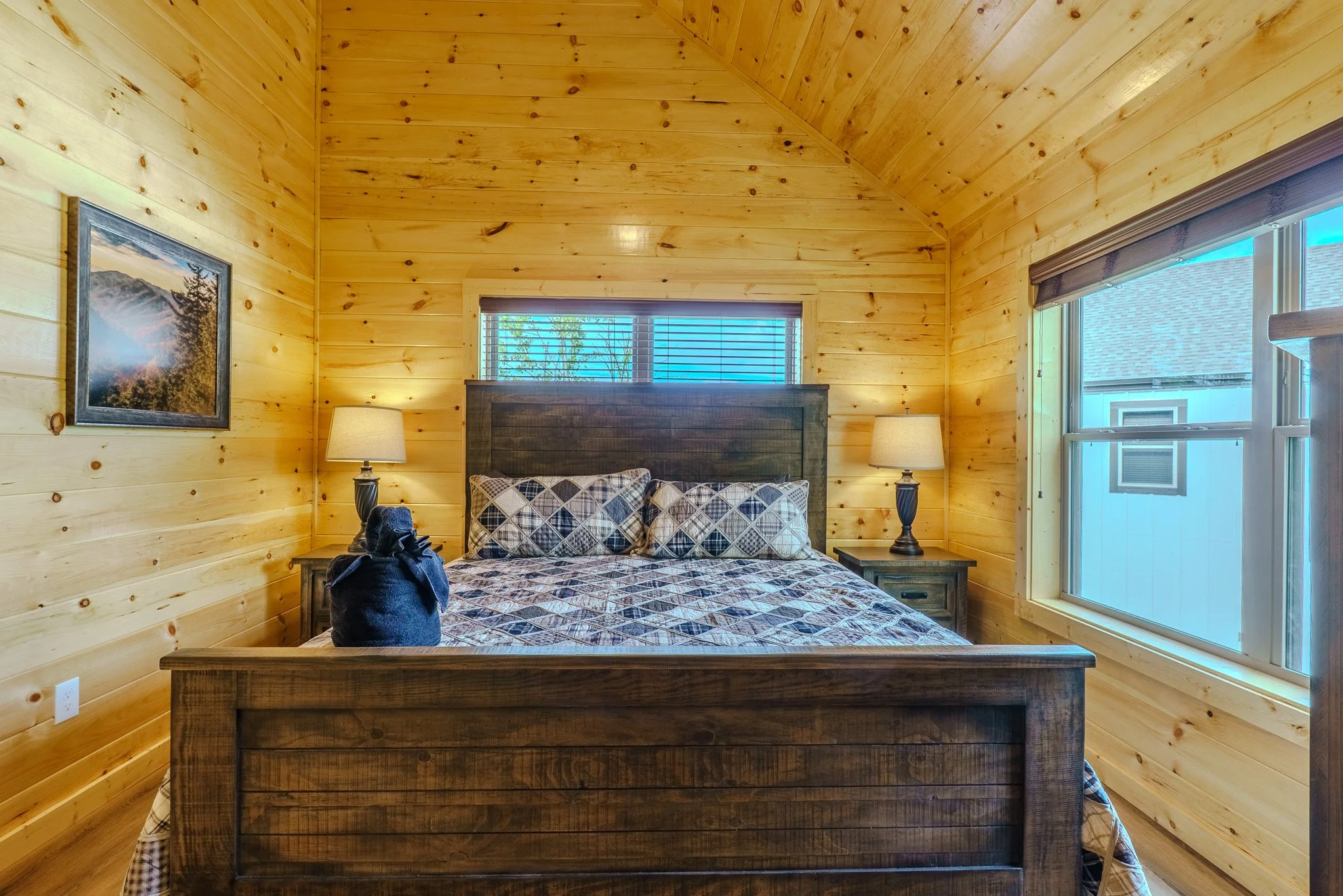Bedroom with wood-paneled walls, a wooden bed with checkered bedding, two nightstands with lamps, a window above the bed, and another window with a view of neighboring houses.